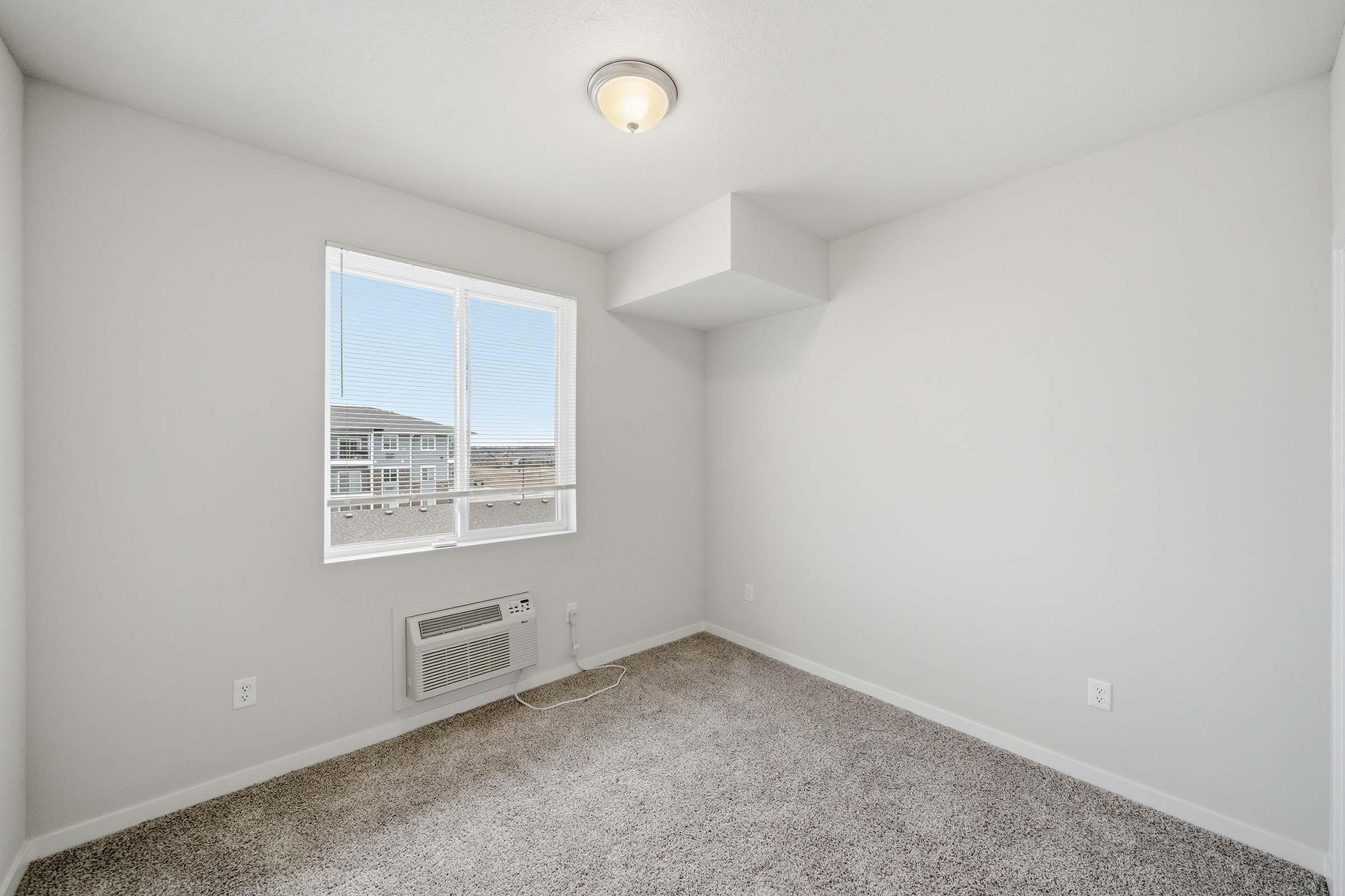 A bright, empty room with light gray walls and carpeted flooring. There is a single ceiling light fixture and a window allowing natural light to enter, showing a view outside. An air conditioning unit is installed beneath the window. The overall space appears clean and well-maintained.