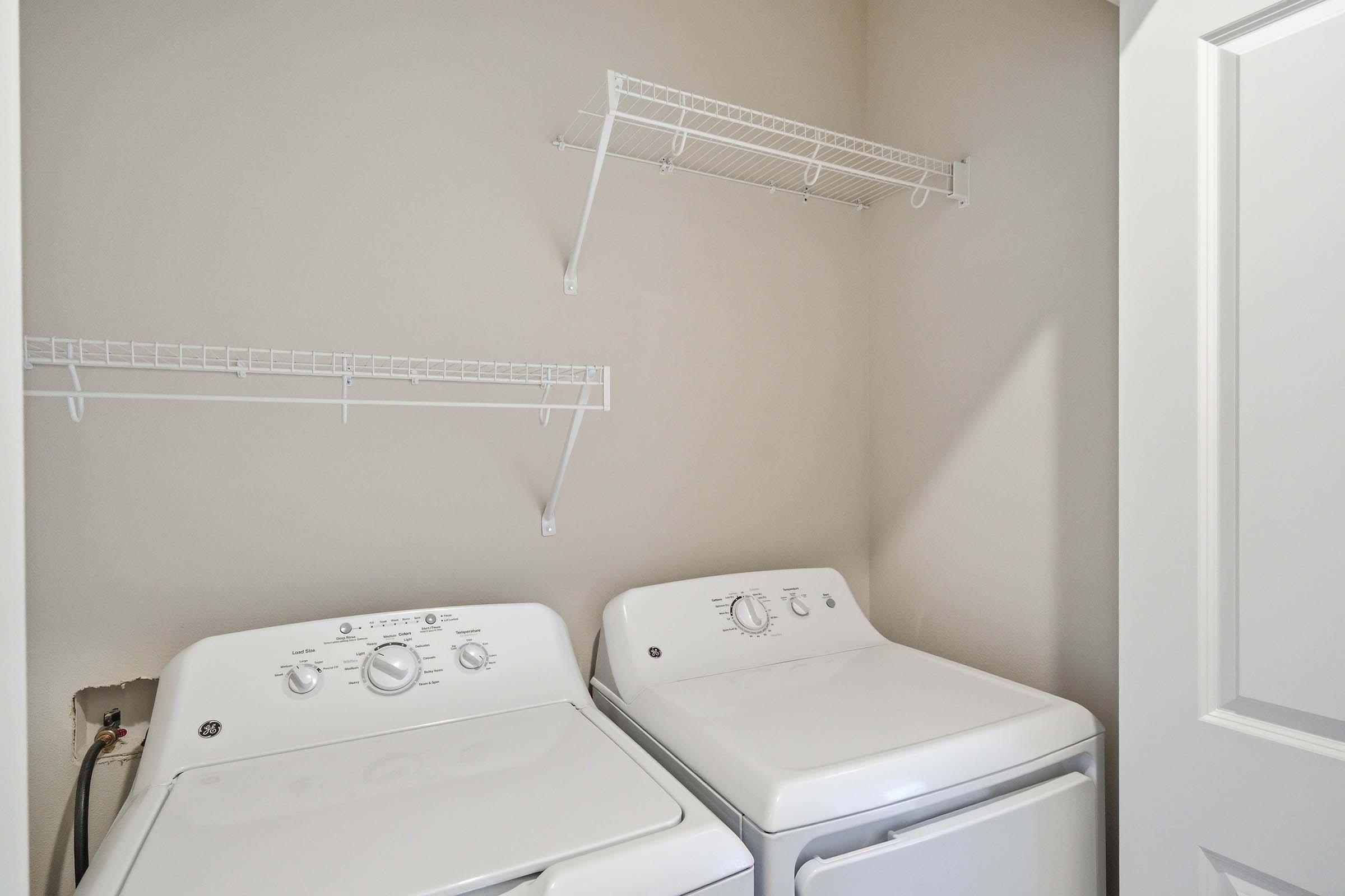A laundry room featuring a stacked washer and dryer side by side. Above them, there are two wire shelving units mounted on the wall. The walls are painted a neutral color, and a door is partially visible to the right.