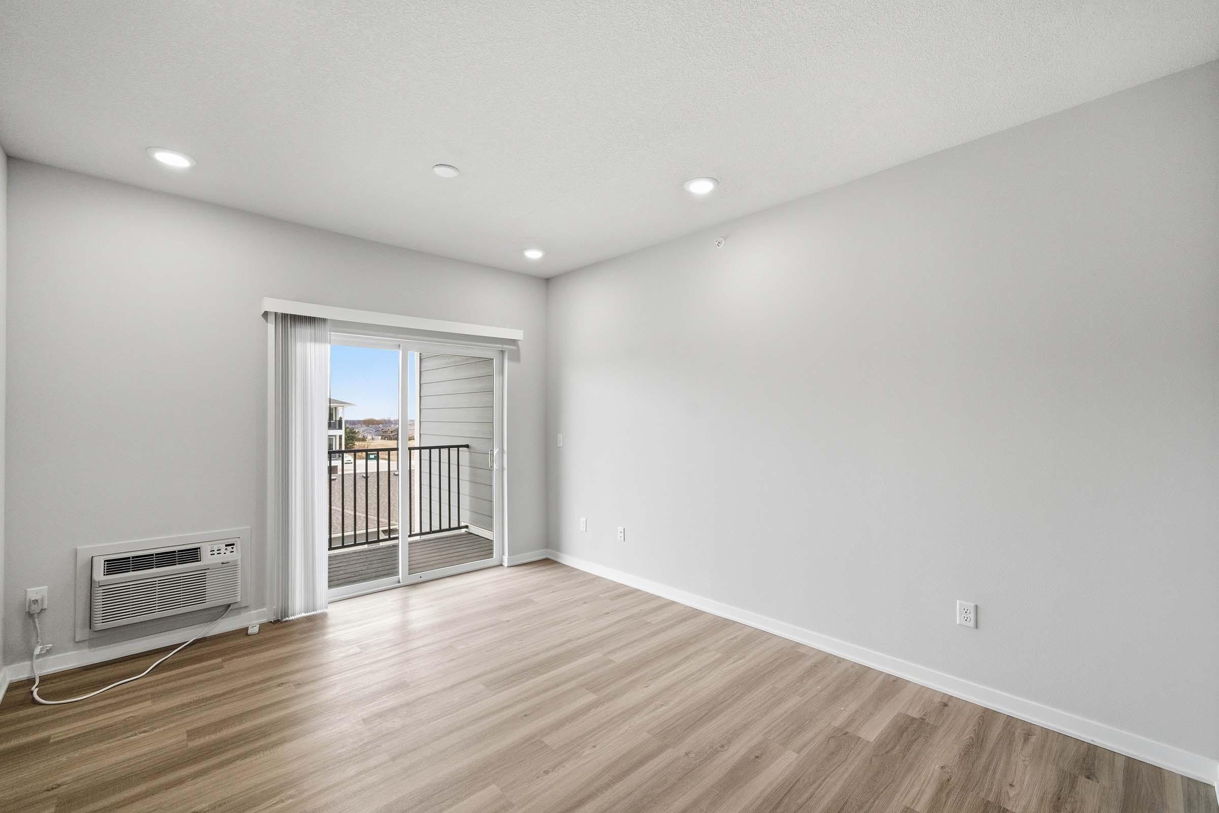 Bright and spacious living room featuring light-colored walls, large windows with a balcony view, and wooden flooring. Equipped with modern lighting and an air conditioning unit. Ideal for creating a comfortable living space.