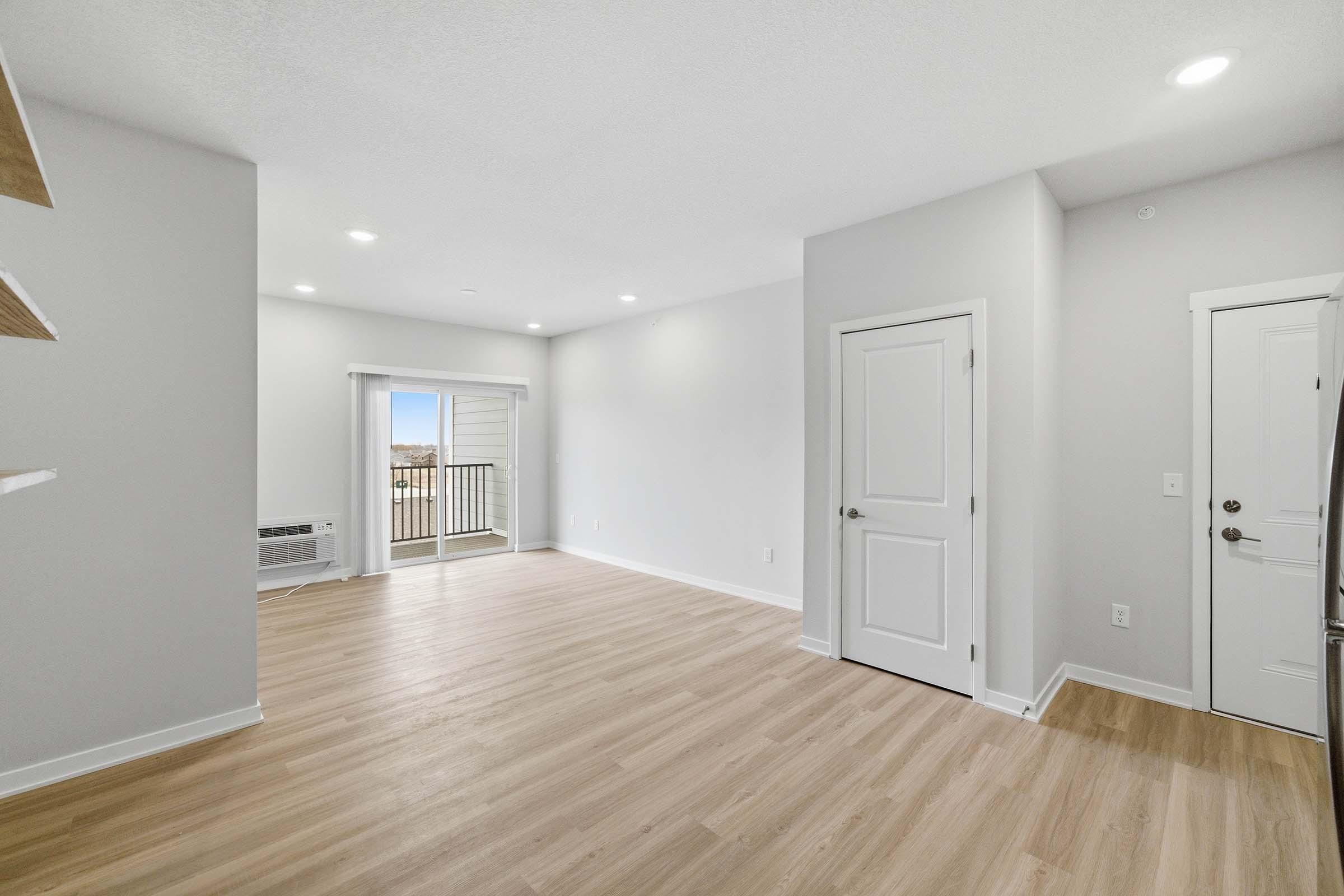 Spacious interior of a modern apartment featuring light-colored walls and flooring, with a door leading to a balcony. Natural light floods the space, showcasing an open layout ideal for living and entertainment. Minimalist design elements create a fresh and inviting atmosphere.