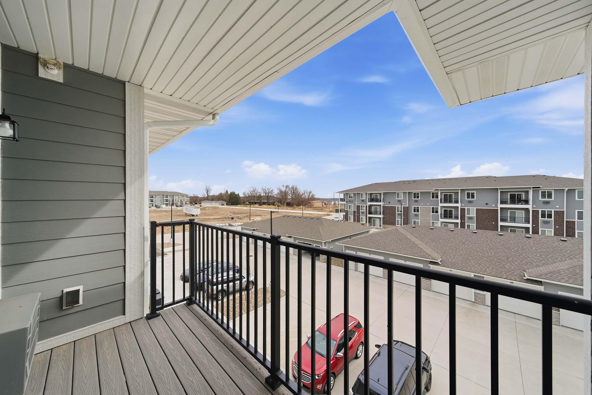 View from a balcony overlooking a parking area and nearby buildings under a clear blue sky. The image features a wooden deck, metal railing, and exterior of a gray building, with cars parked below. The landscape is mostly vacant with a few structures in the distance.
