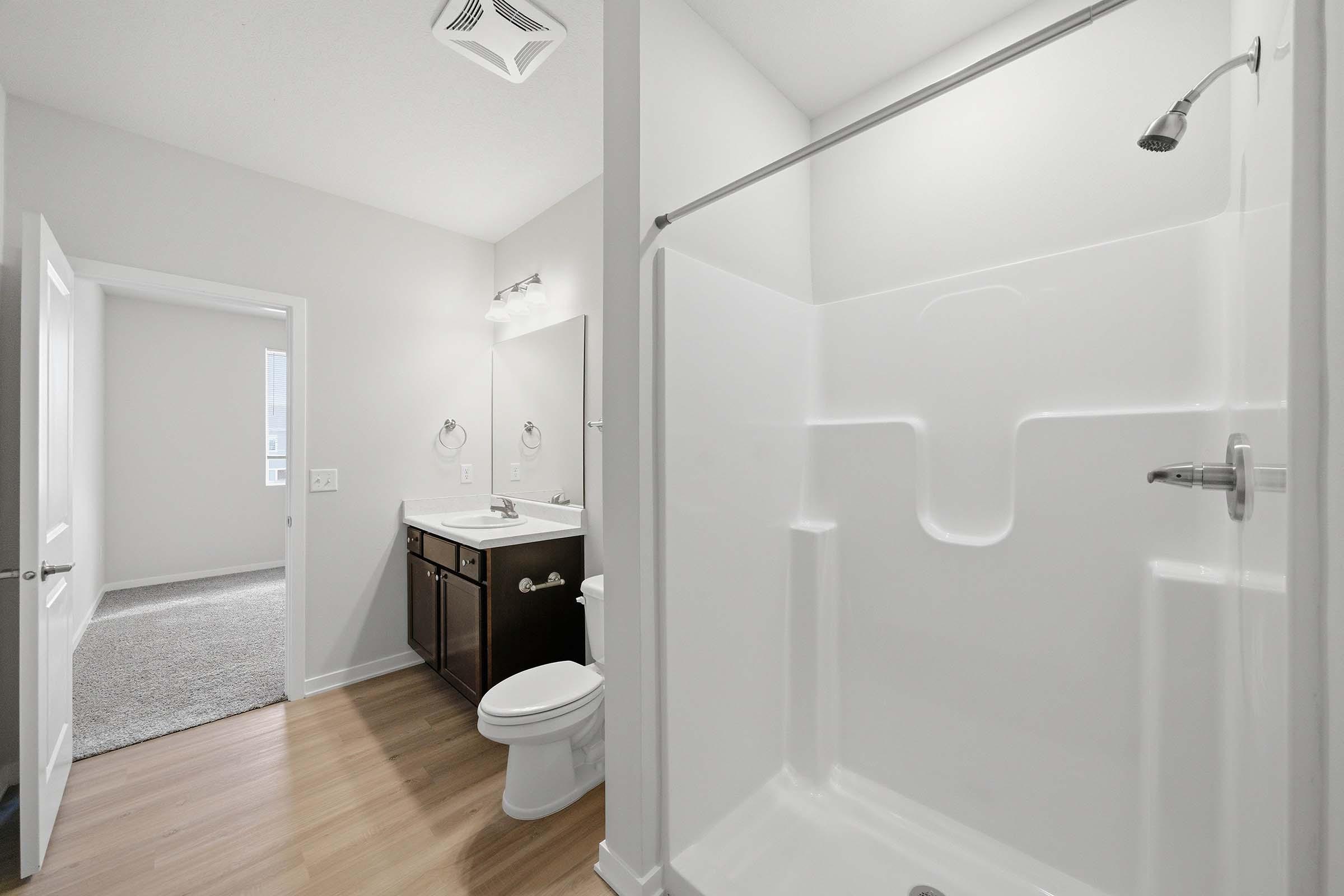 A modern bathroom featuring a white shower stall, a sink with a dark wood vanity, and a toilet. The walls are light-colored, and there is a mirror above the sink. A doorway leads to another room, and the floor is made of wood-like tiles. Natural light enters through a window in the adjacent room.