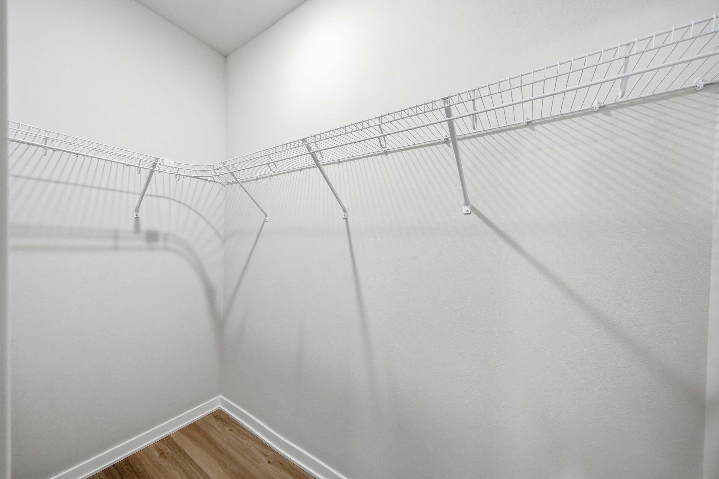 An empty walk-in closet with white wire shelving against light-colored walls and wooden flooring. The shelves are arranged in a corner, casting shadows on the wall. There are no items or clothing visible in the closet.