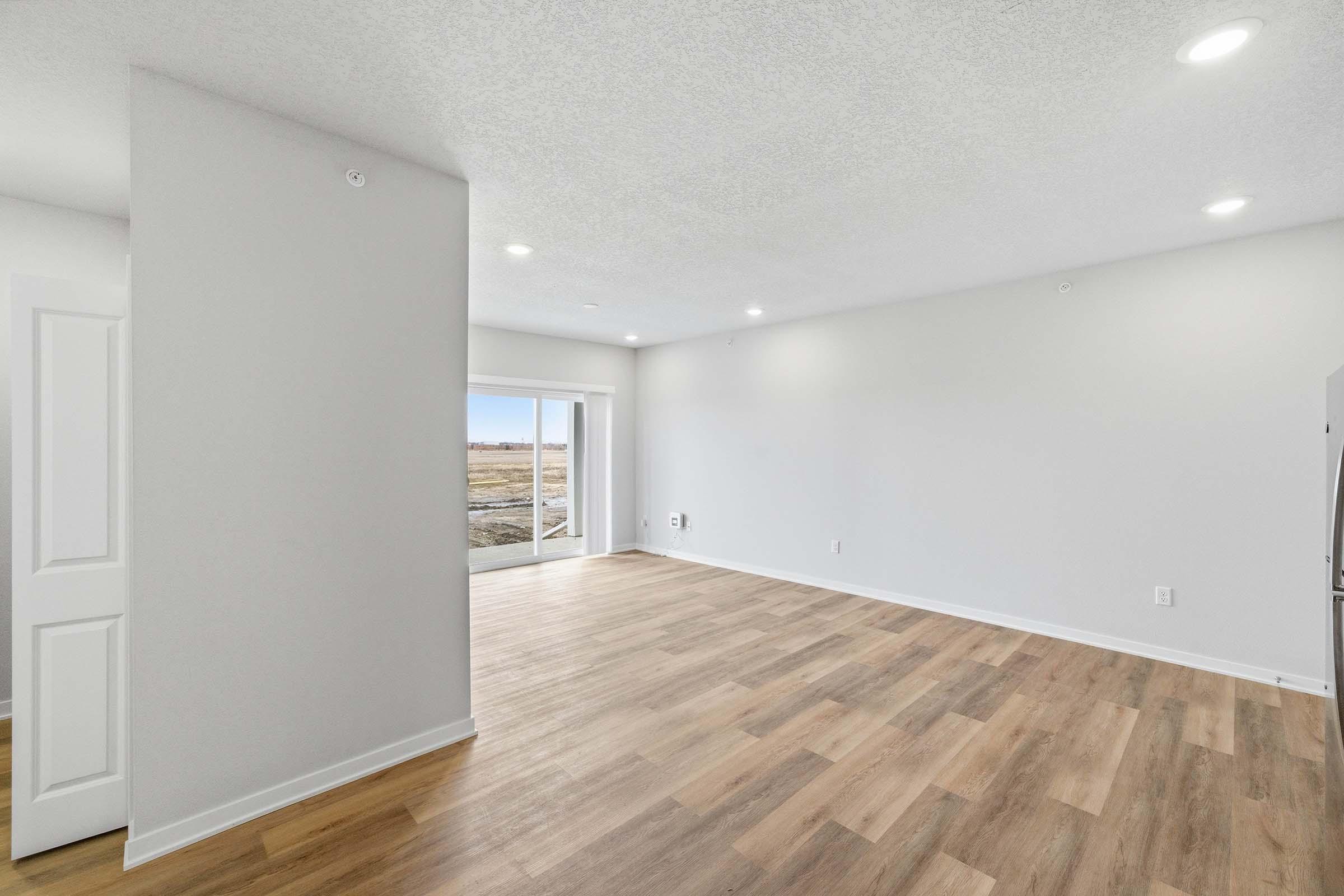 A spacious, empty room with light-colored walls and a textured ceiling. The floor is made of light wood laminate. A large sliding glass door at one end opens to a view of an outdoor space, with natural light illuminating the area. The room is unfurnished, creating a clean and minimalist look.