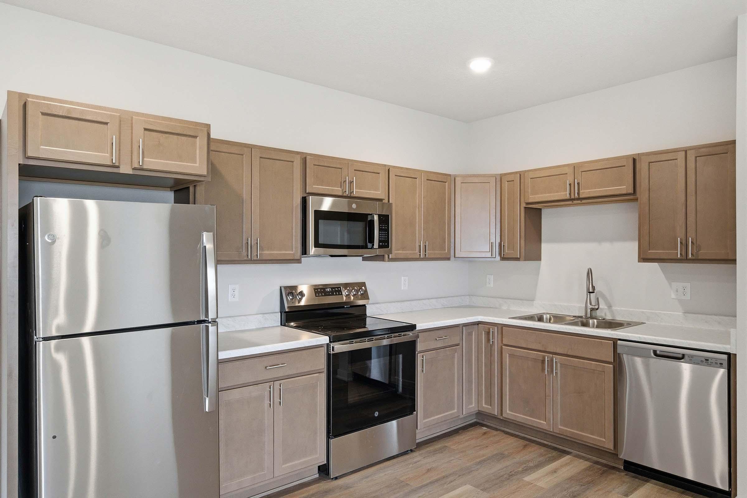 Modern kitchen featuring stainless steel appliances, including a refrigerator, microwave, and stove. The cabinetry is a light wood finish, and the countertops are a clean, light color. There's a sink next to the dishwasher, and the flooring is light-colored wood. Brightly lit with recessed lighting.