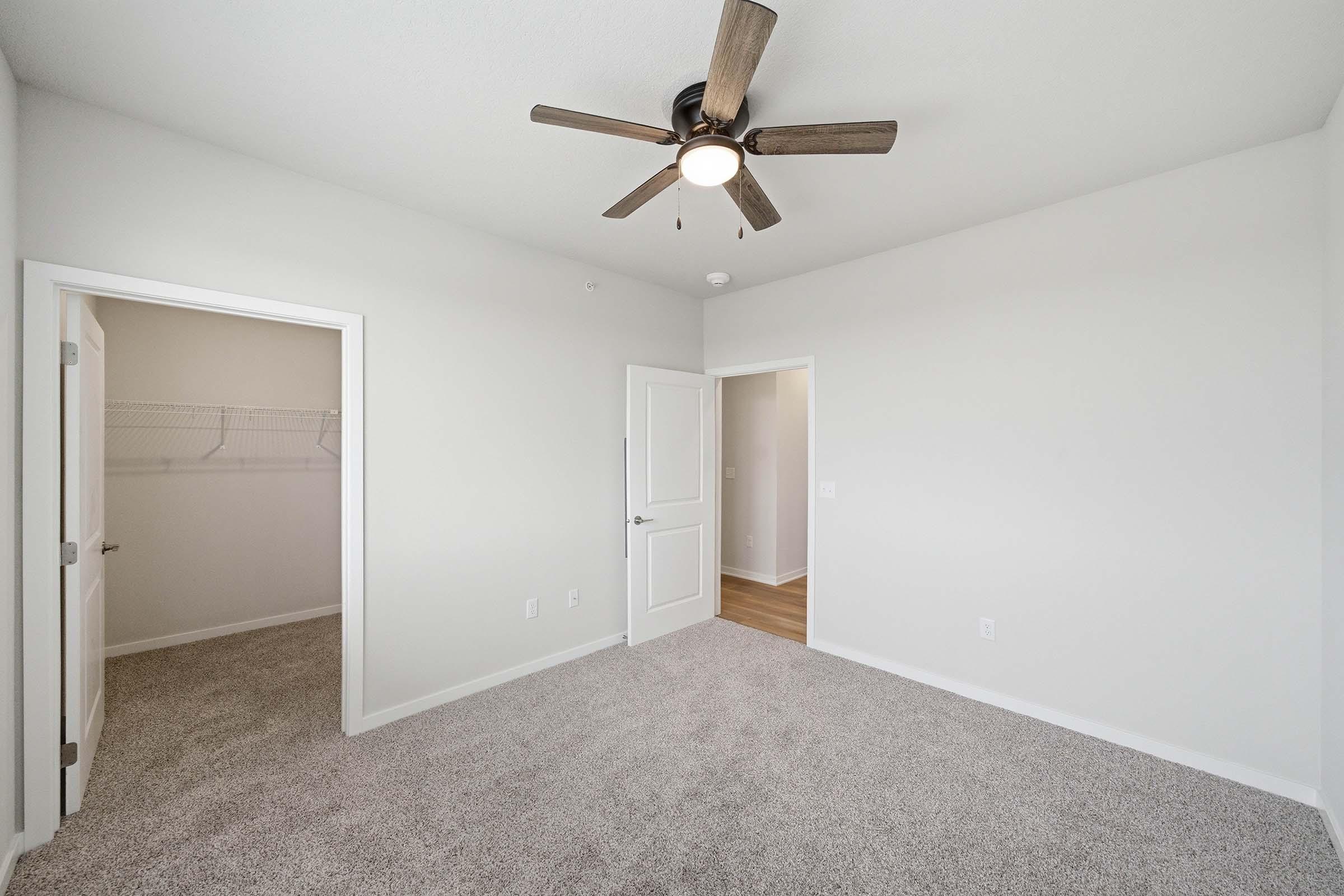 A well-lit, empty bedroom featuring light-colored walls and a ceiling fan. There is plush carpeting on the floor and two doorways leading to a closet and an adjacent room, enhancing the sense of space and openness.