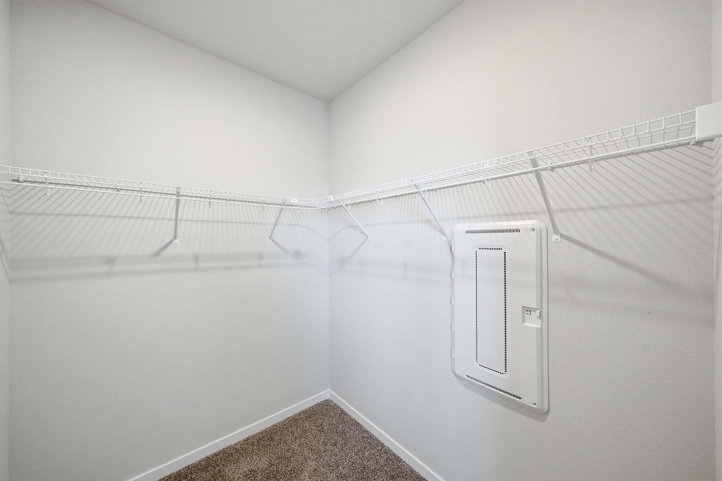 A spacious, empty closet featuring white wire shelving mounted on light gray walls. The floor is carpeted in a neutral tone, and there is a small panel or door on one wall, possibly for utility access. The overall look is clean and minimalistic, providing a blank canvas for storage.