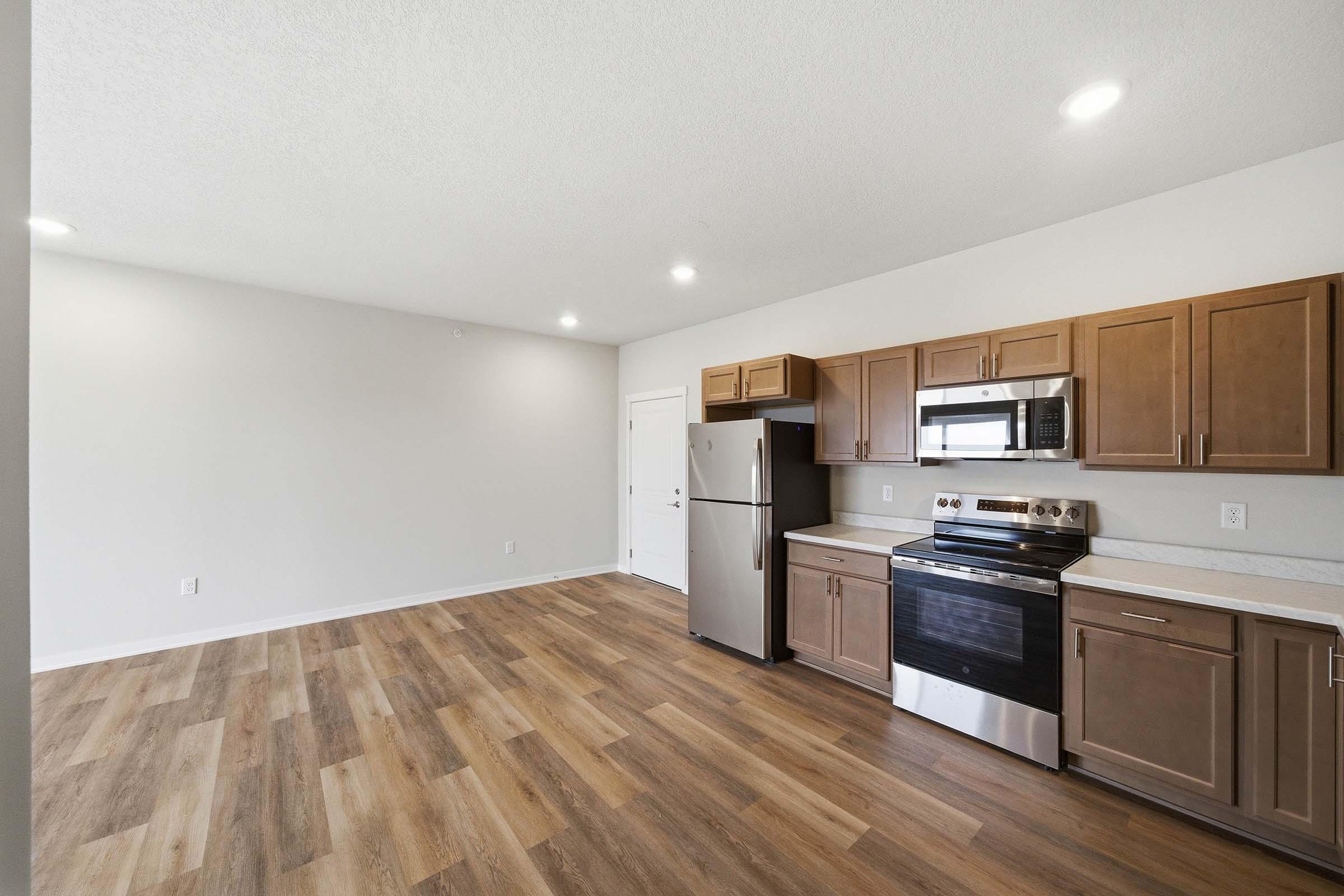 Modern kitchen with stainless steel appliances, including a refrigerator and oven, wooden cabinetry, and a light-colored countertop. The space features an open layout with wood-style flooring and neutral-colored walls, creating a bright and inviting atmosphere.