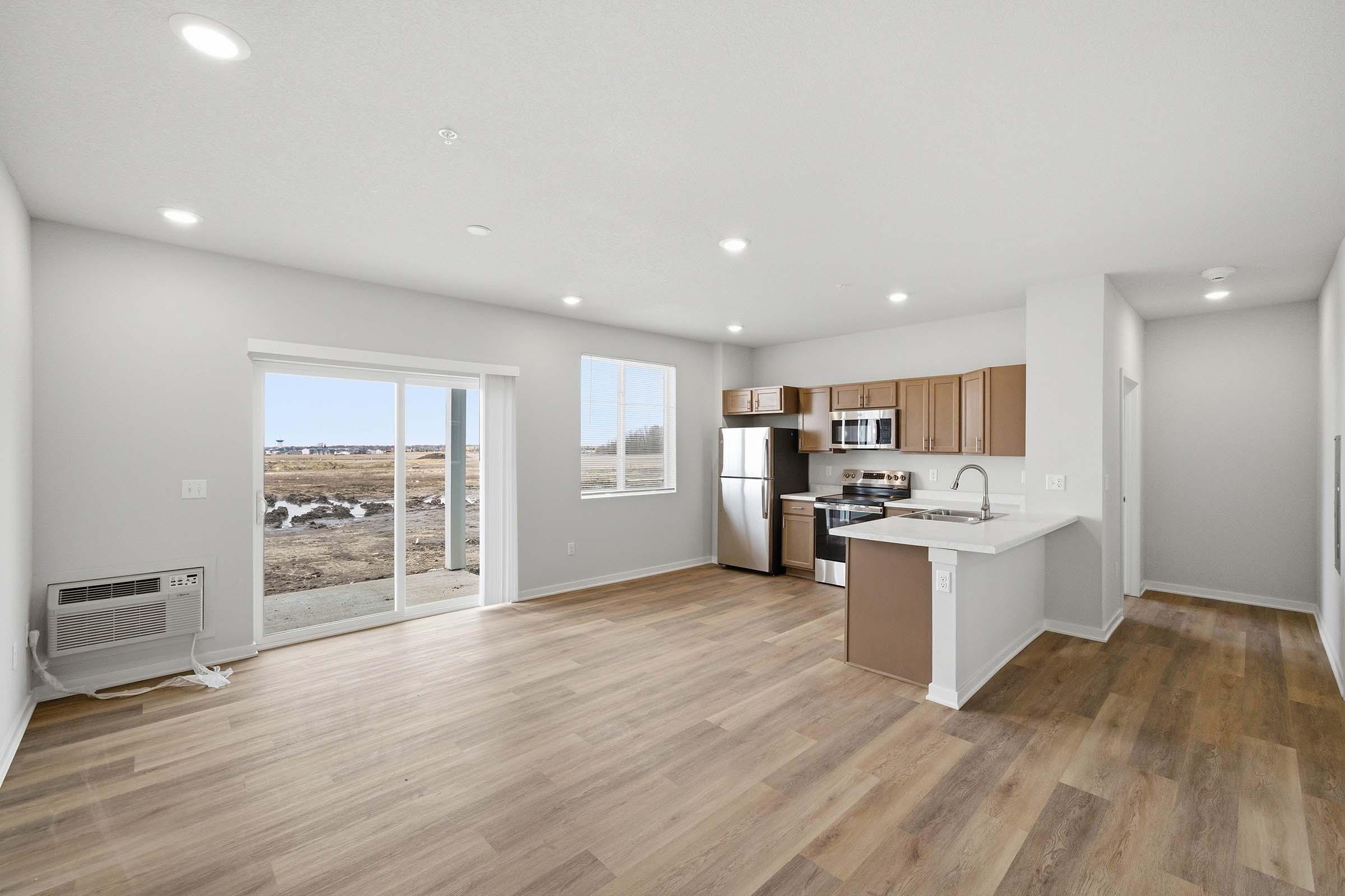 Bright and open interior of a modern kitchen and living area, featuring light wood flooring, large windows with views outside, a sliding glass door, stainless steel appliances, and wooden cabinets. The layout is spacious and inviting, emphasizing natural light and a contemporary design.