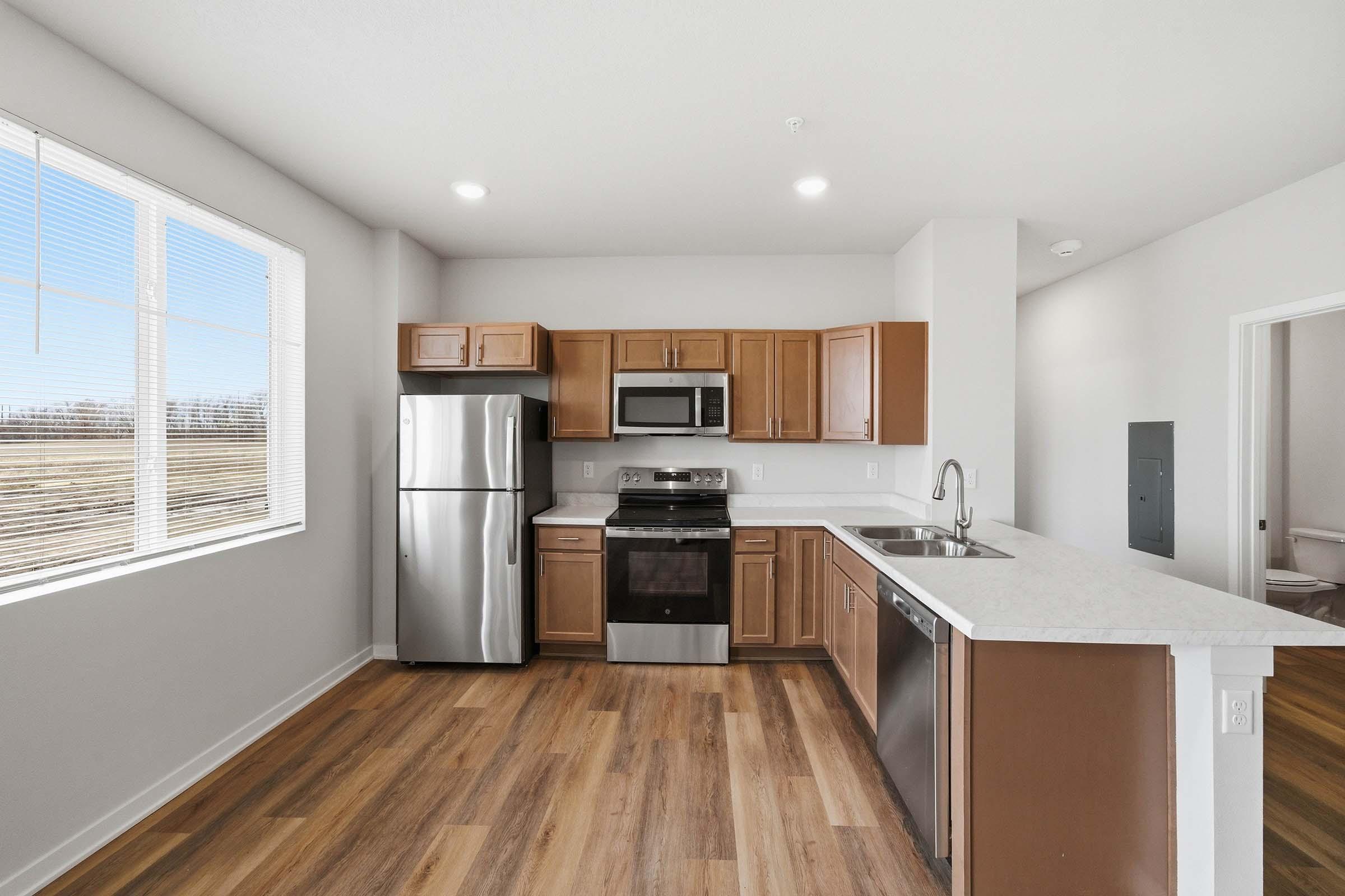 Modern kitchen featuring stainless steel appliances including a refrigerator, oven, and microwave. The cabinetry is a warm wood tone, and the countertops are light-colored. Large window provides natural light, and there are wood-style floors. The layout is open and spacious, with a countertop for additional seating.