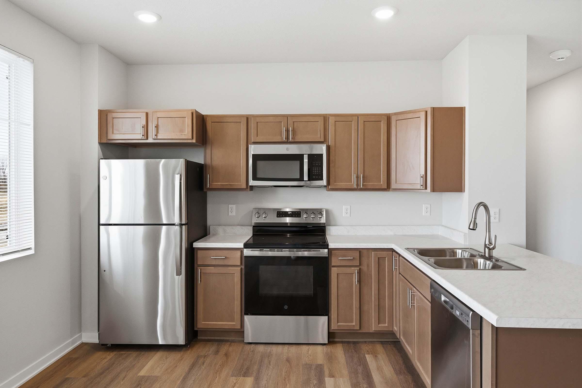 Modern kitchen featuring stainless steel appliances, including a refrigerator, microwave, and oven. The cabinets are a warm wood tone, and the countertops are white with a subtle pattern. Natural light comes through the window, illuminating the space, which has a clean and contemporary design.