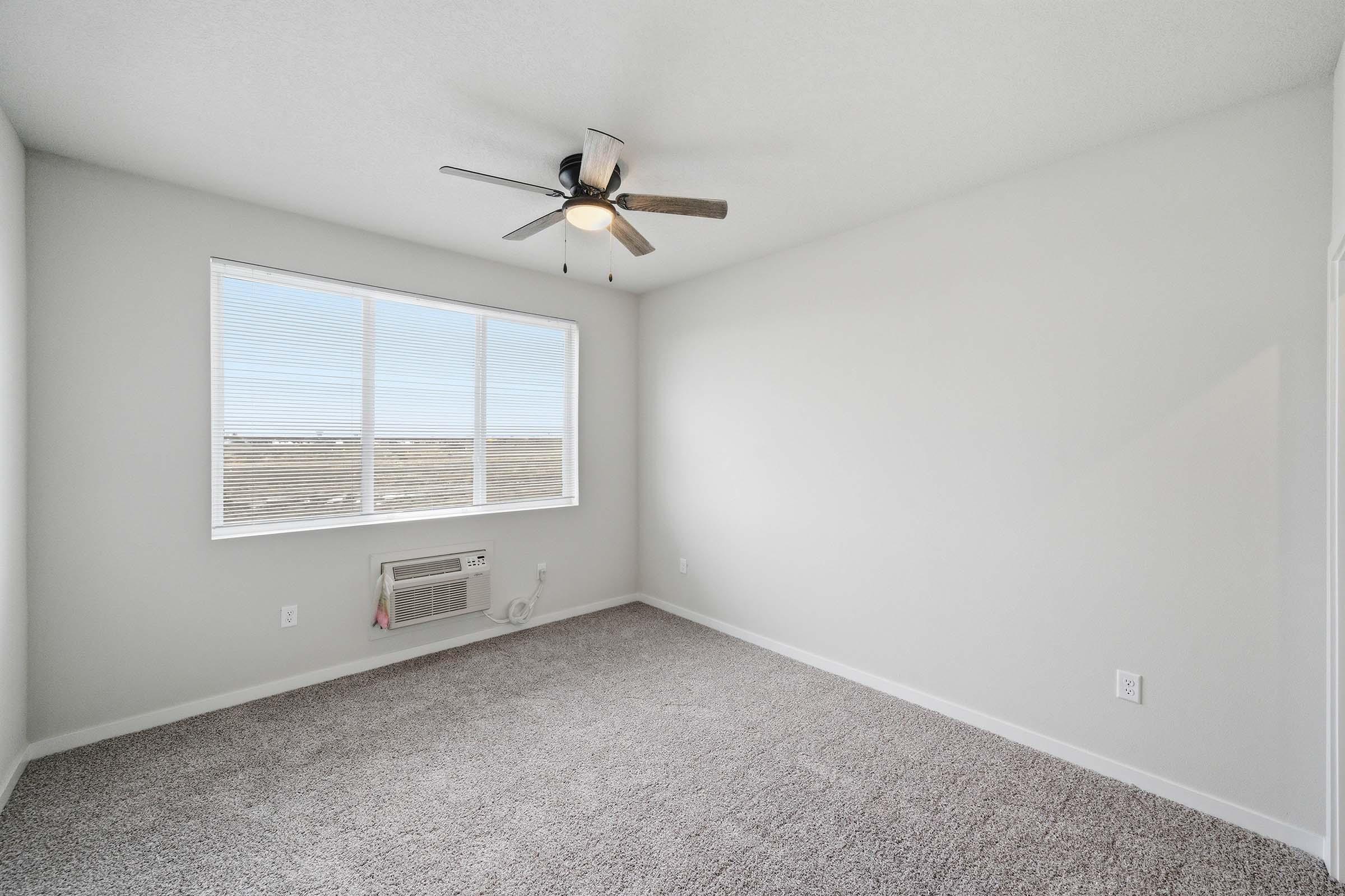 A bright, empty room with light gray walls and a light-colored carpet. A ceiling fan with wooden blades hangs from the ceiling. A large window lets in natural light, showcasing a view outside. An air conditioning unit is mounted beneath the window. The room has a minimalist and spacious feel.