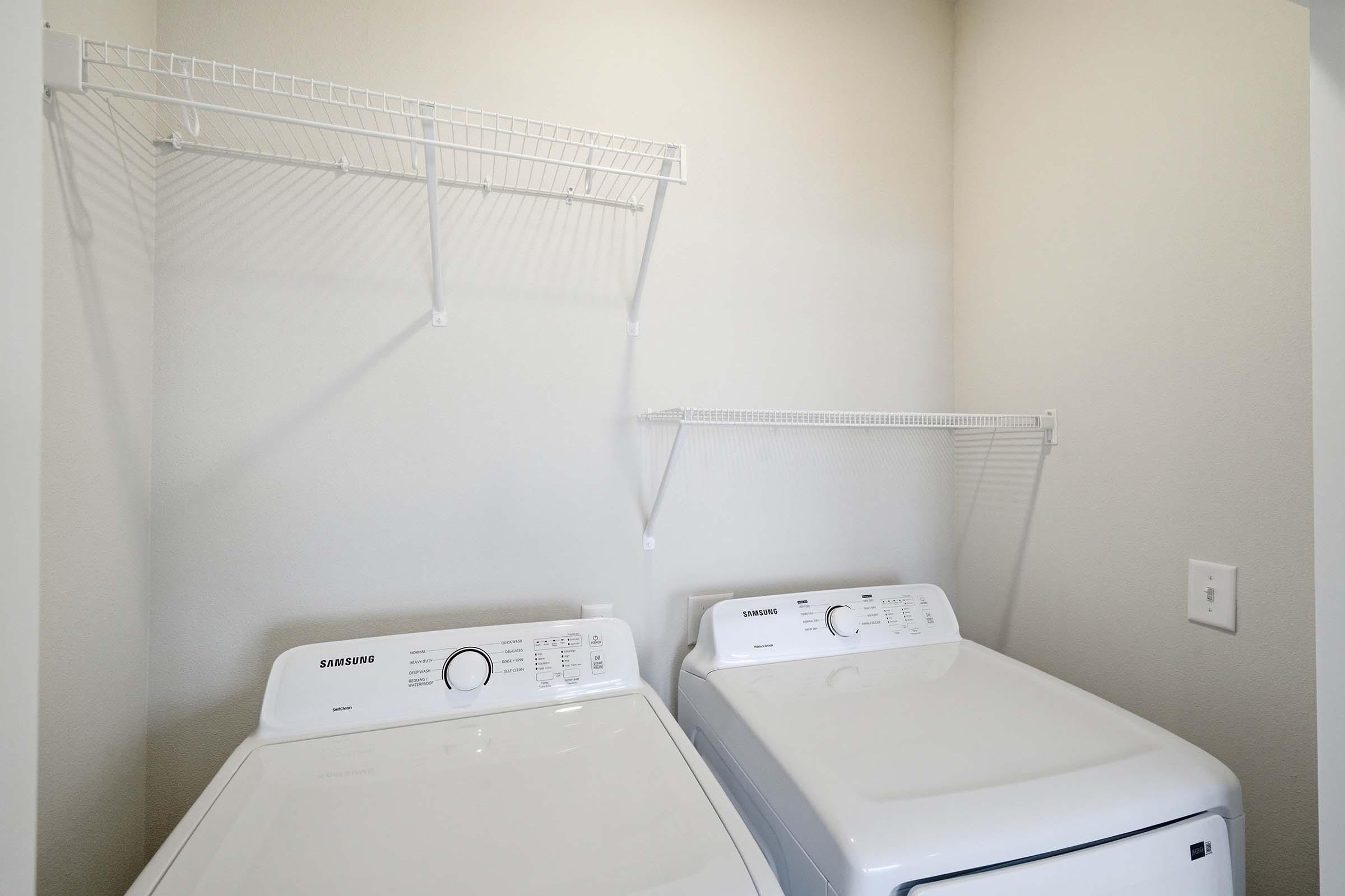 A laundry room featuring a washing machine and a dryer. Both appliances are white and stacked side by side. Above them, there is a wire shelf for storage. The wall is painted in a light color, and there is an electrical outlet visible nearby.