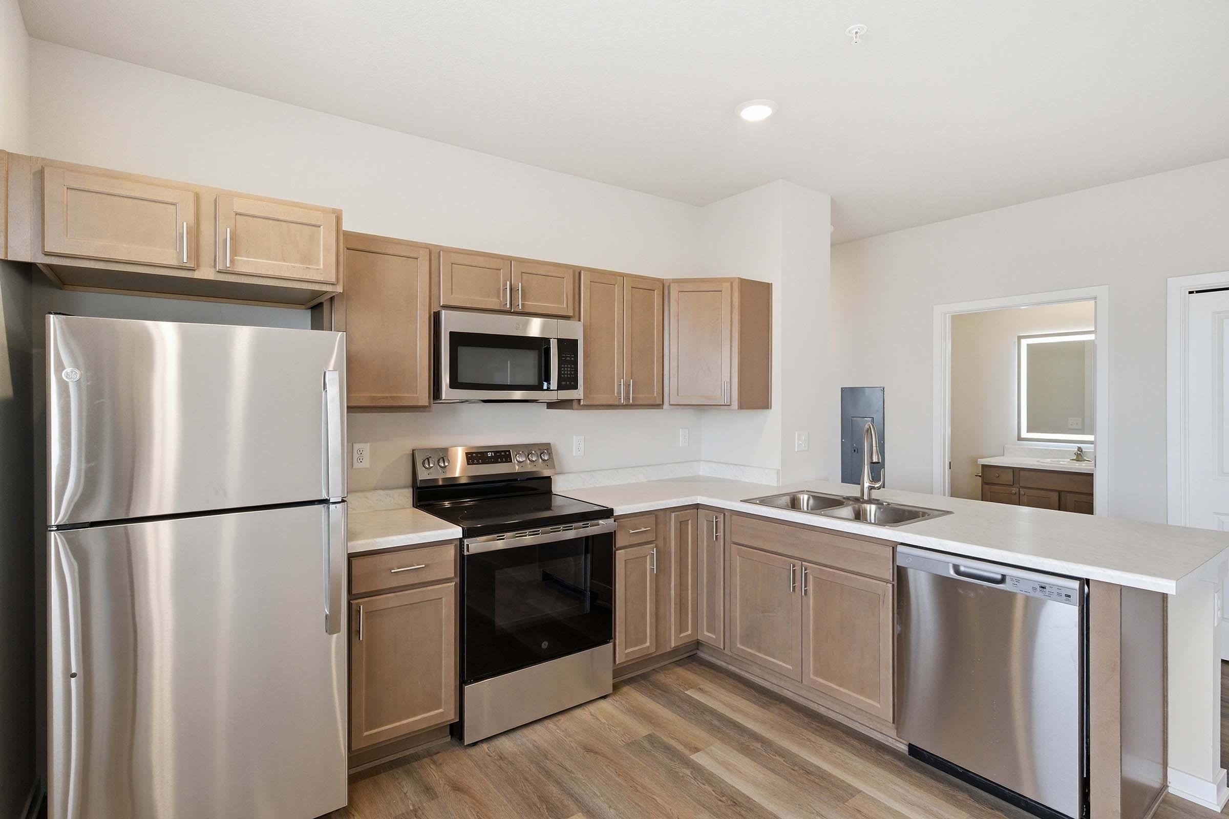 A modern kitchen featuring light wooden cabinetry, stainless steel appliances including a refrigerator, microwave, and oven. The countertop is a light color, and there's a double sink. The kitchen has a neutral color scheme and is well-lit, with a small mirror visible in an adjoining space.