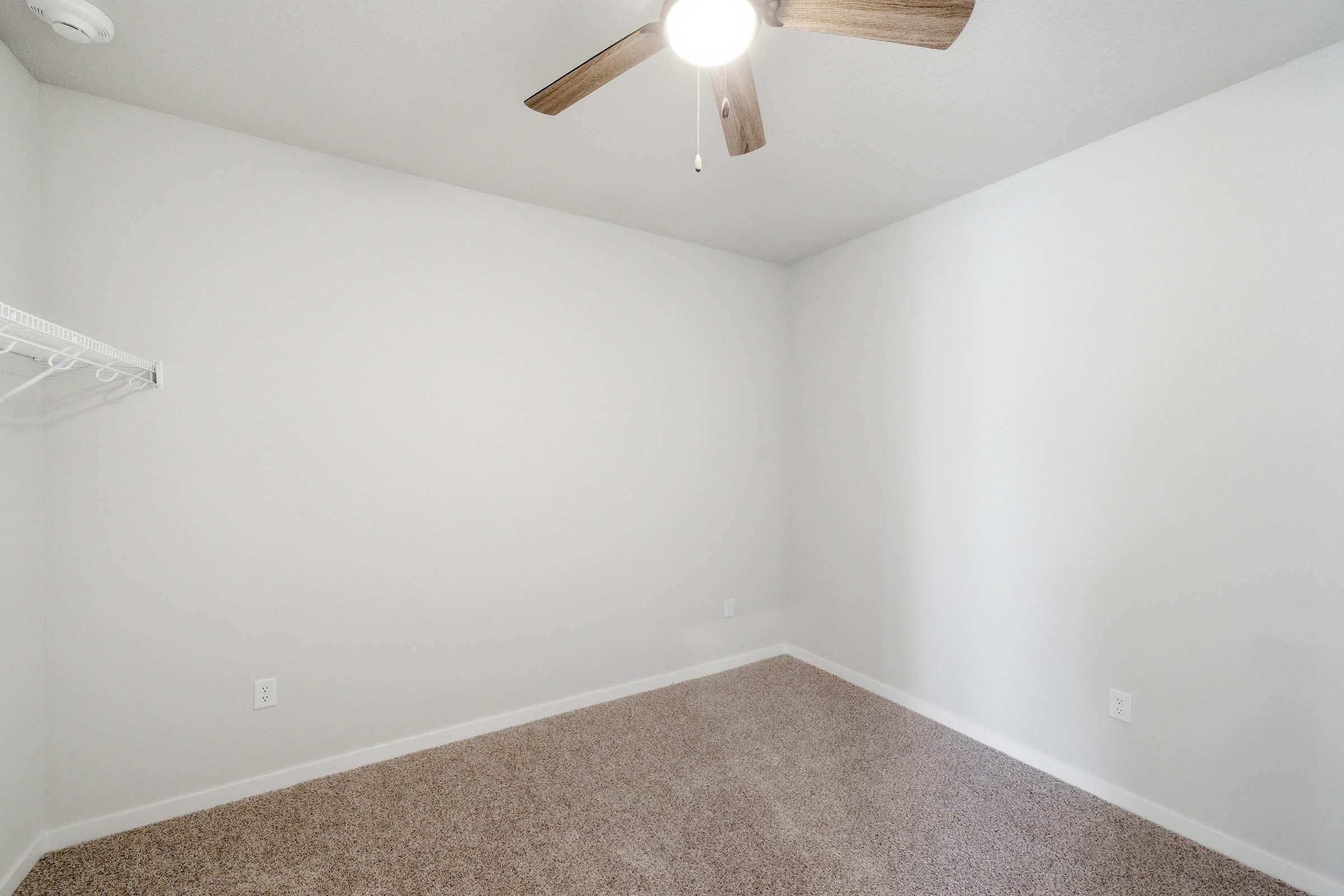 A vacant room with light-colored walls and a carpeted floor. There's a ceiling fan with wooden blades, and a wall-mounted shelf on the left. The room appears bright and spacious, suitable for various uses. The overall atmosphere is clean and minimalistic.