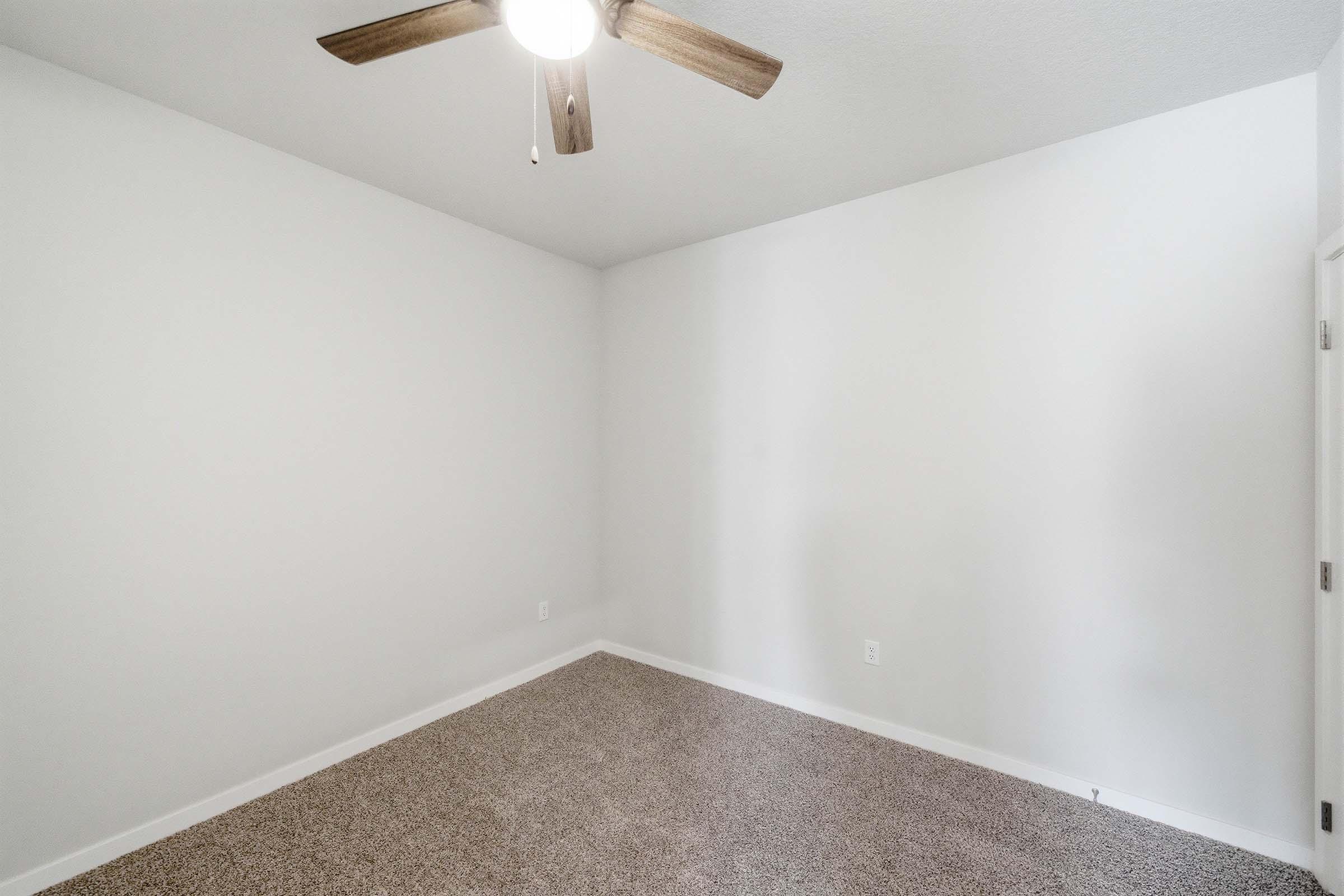 Empty room with light gray walls and a ceiling fan. The floor is covered in beige carpet. There is a single door on the right side of the image, and the room appears bright and spacious with no furniture or decorations.