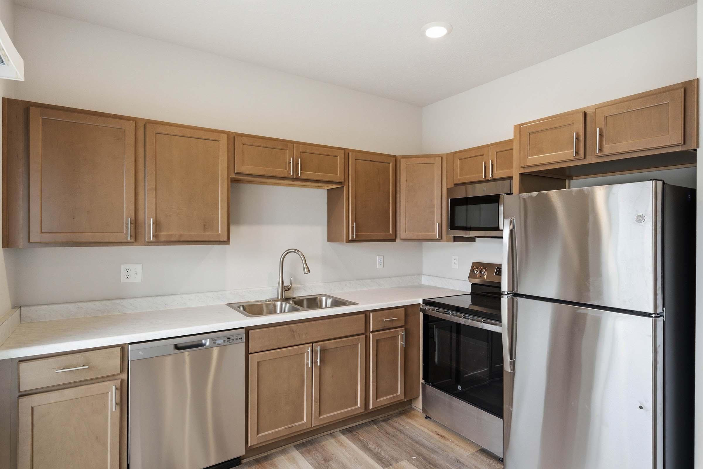 A modern kitchen featuring light brown wooden cabinets, a stainless steel refrigerator, and a dishwasher. The countertop is light-colored, and there is a sink with a single faucet. Appliances include a microwave and an oven with a stovetop. The kitchen has a bright, clean aesthetic.
