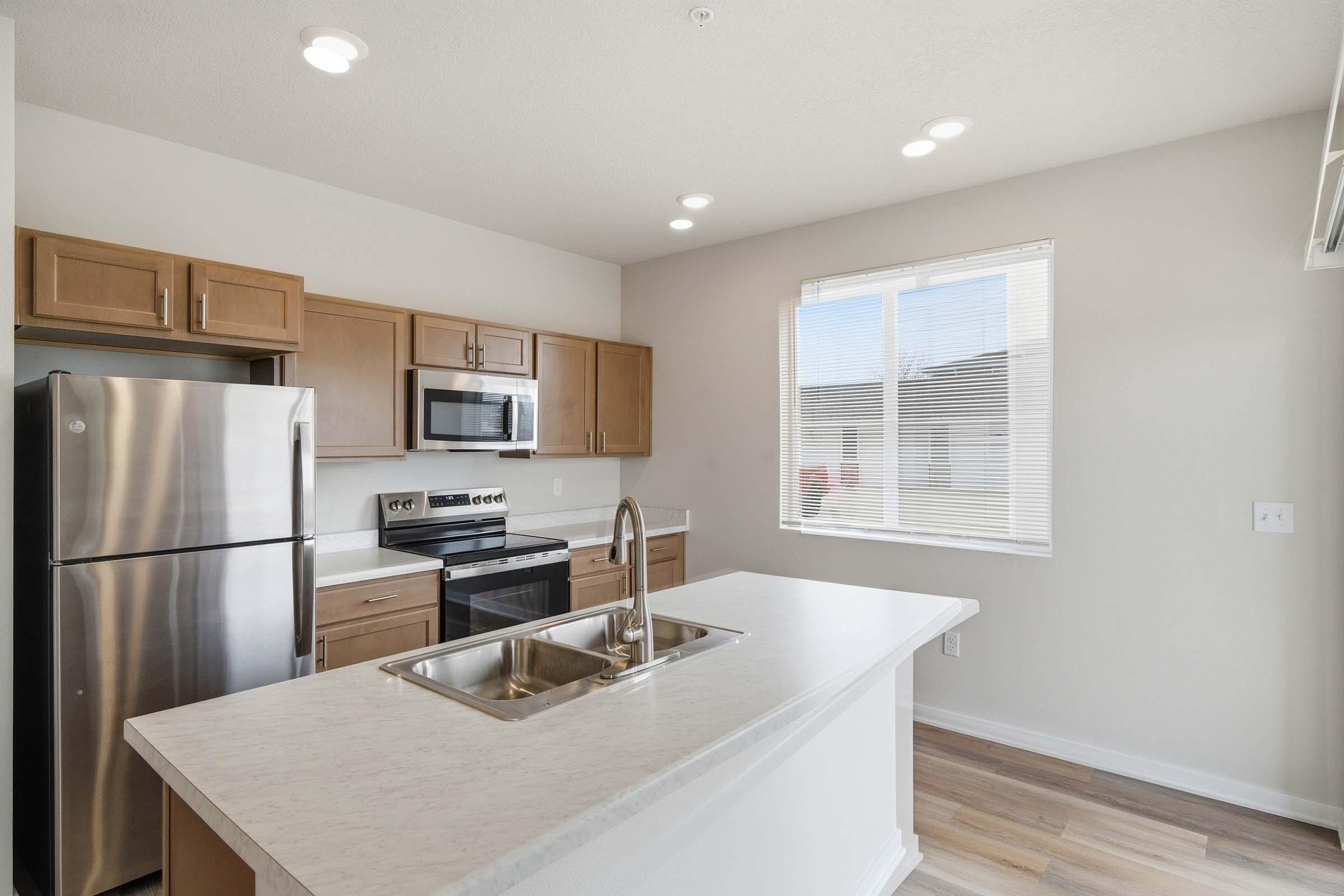 Modern kitchen featuring stainless steel appliances, including a refrigerator and microwave, along with wooden cabinetry. A large island with a double sink sits in the center, complemented by ample natural light coming through a nearby window. Neutral colors enhance the spacious feel.