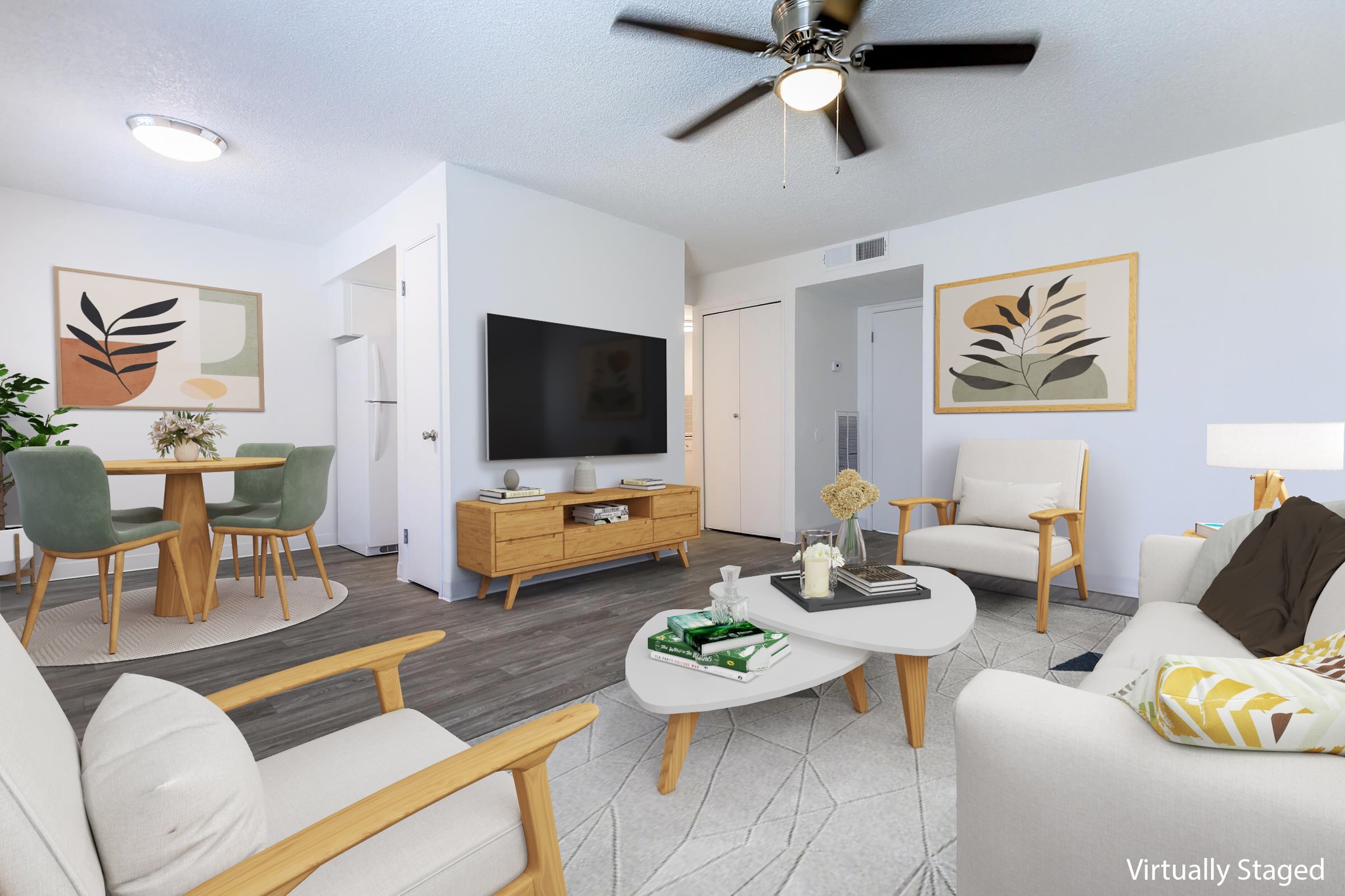 Modern living room showcasing a cozy layout. Features include two armchairs, a sofa, and a coffee table. A wall-mounted TV is on a wooden media console. Decorative plants and abstract wall art enhance the space. A dining area with a round table and green chairs is visible in the background. Bright and inviting atmosphere.