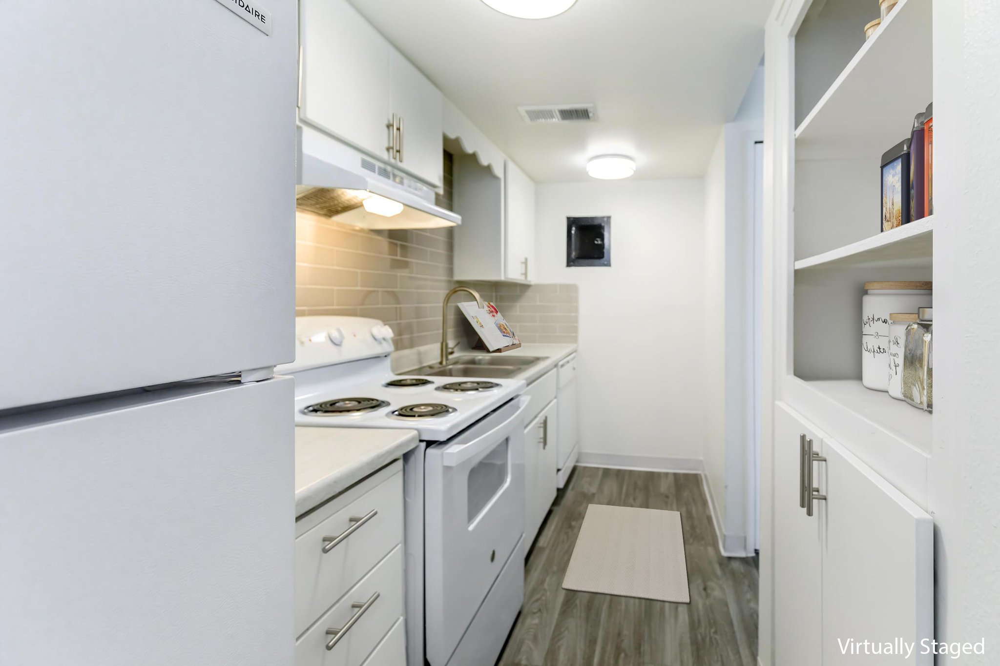 Narrow kitchen with white cabinets and appliances, including a refrigerator and oven. The countertops are light-colored, and there's a small rug on the floor. A shelf is visible on the right, holding various items. Soft overhead lighting illuminates the space.