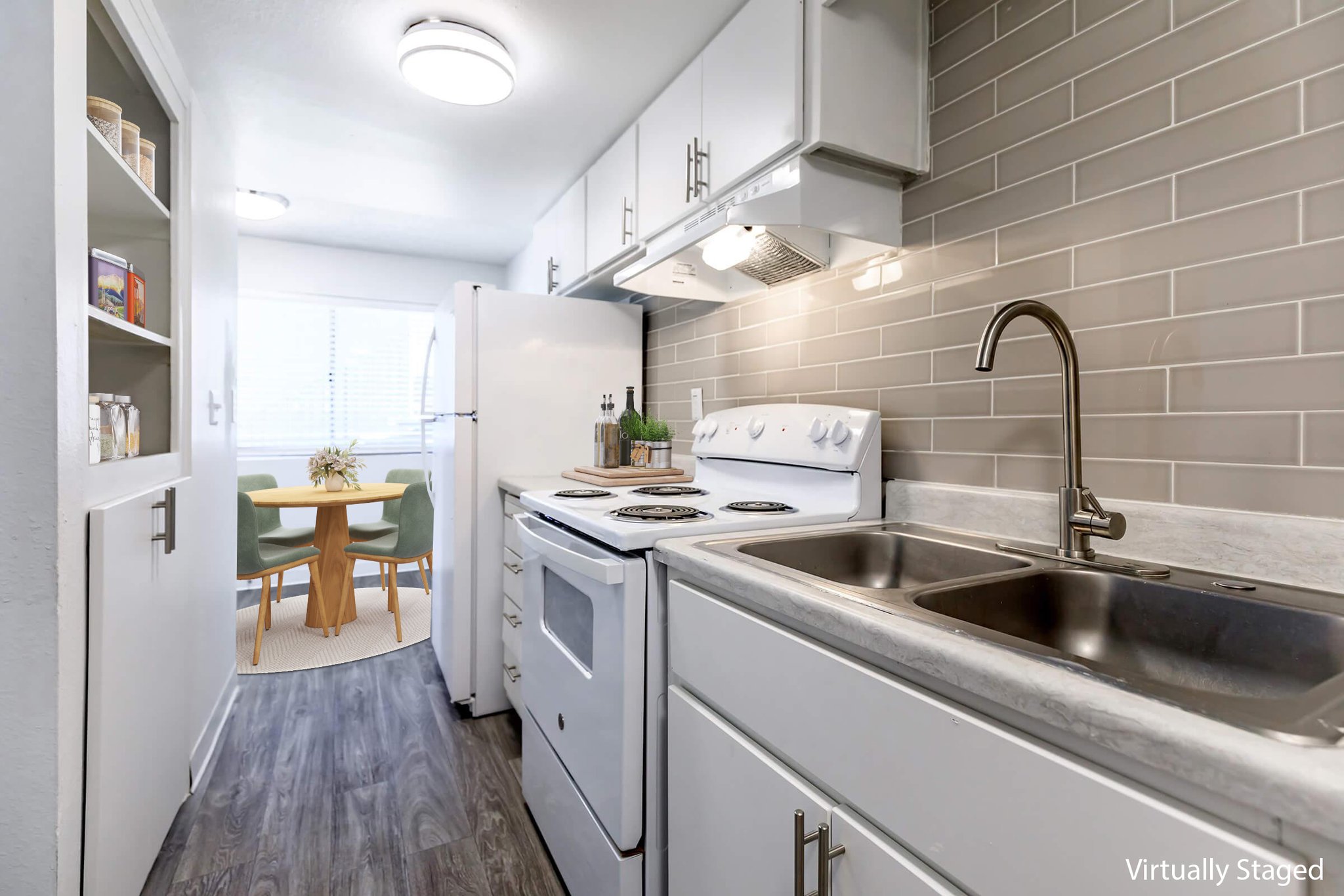 A modern kitchen featuring white cabinetry, a stainless steel sink, and a white stove. The countertop is light-colored, with a decorative tray on it. The kitchen opens to a dining area with a round table and green chairs, illuminated by natural light from a nearby window. The floor has a wood-like finish.