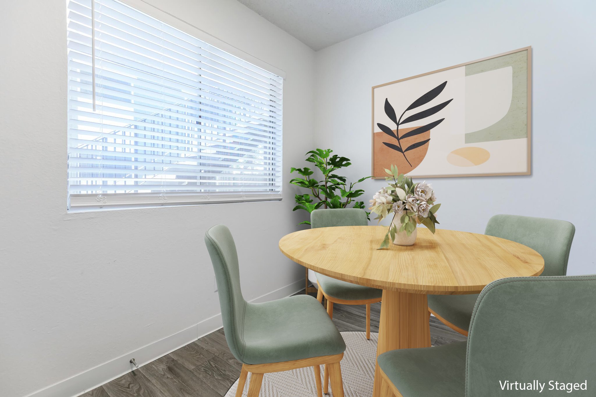 A cozy dining area featuring a round wooden table surrounded by four green chairs. A floral centerpiece sits on the table. The room has a large window with blinds, letting in natural light, and a piece of abstract wall art adorns one wall, complemented by a small potted plant in the corner.