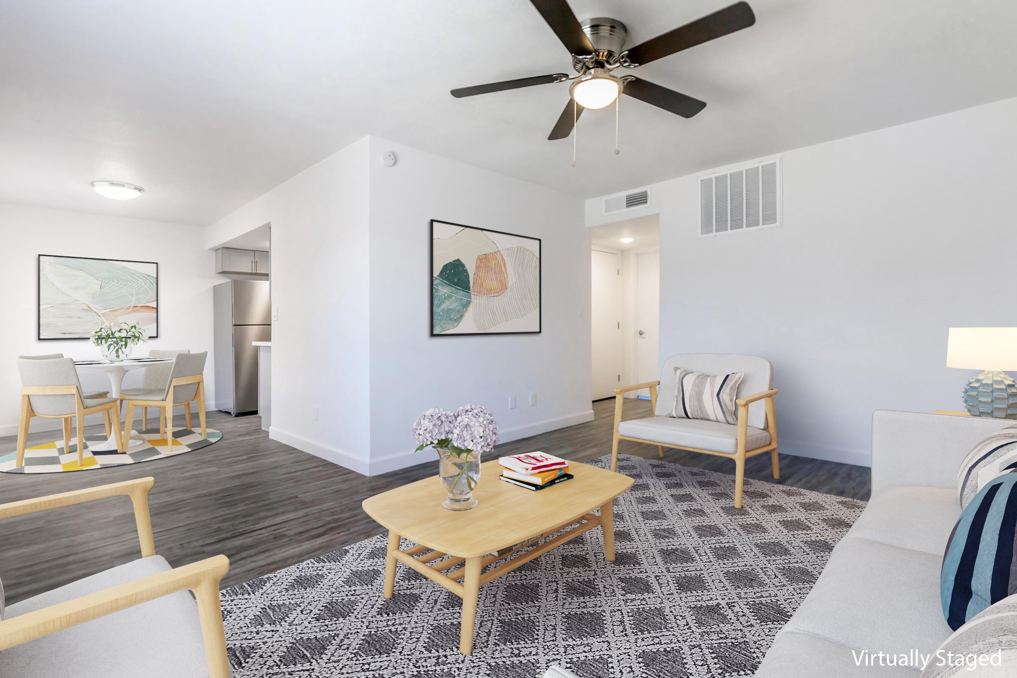 A bright and modern living room featuring a ceiling fan, a beige sofa with decorative pillows, a coffee table with magazines, and a floral centerpiece. Wall art in soft colors adorns the walls. In the background, there's a dining area with a round table and chairs, complemented by a clean, contemporary kitchen space.
