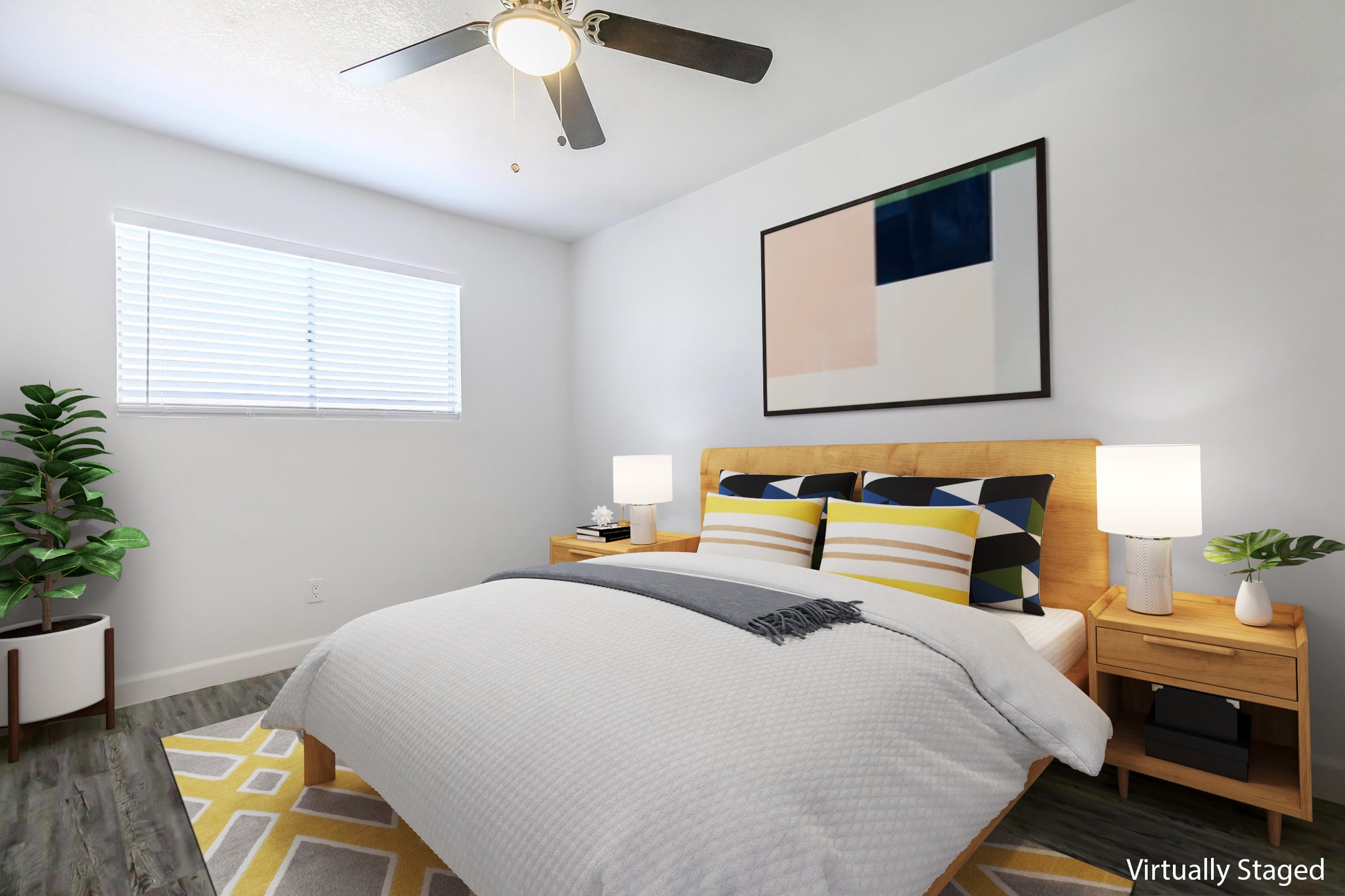 A modern, virtually staged bedroom featuring a queen-sized bed with yellow and blue accent pillows. There are two bedside tables with lamps, a framed abstract wall art piece above the bed, and a plant in the corner. A window with blinds allows natural light into the room. The floor is a light wooden finish.