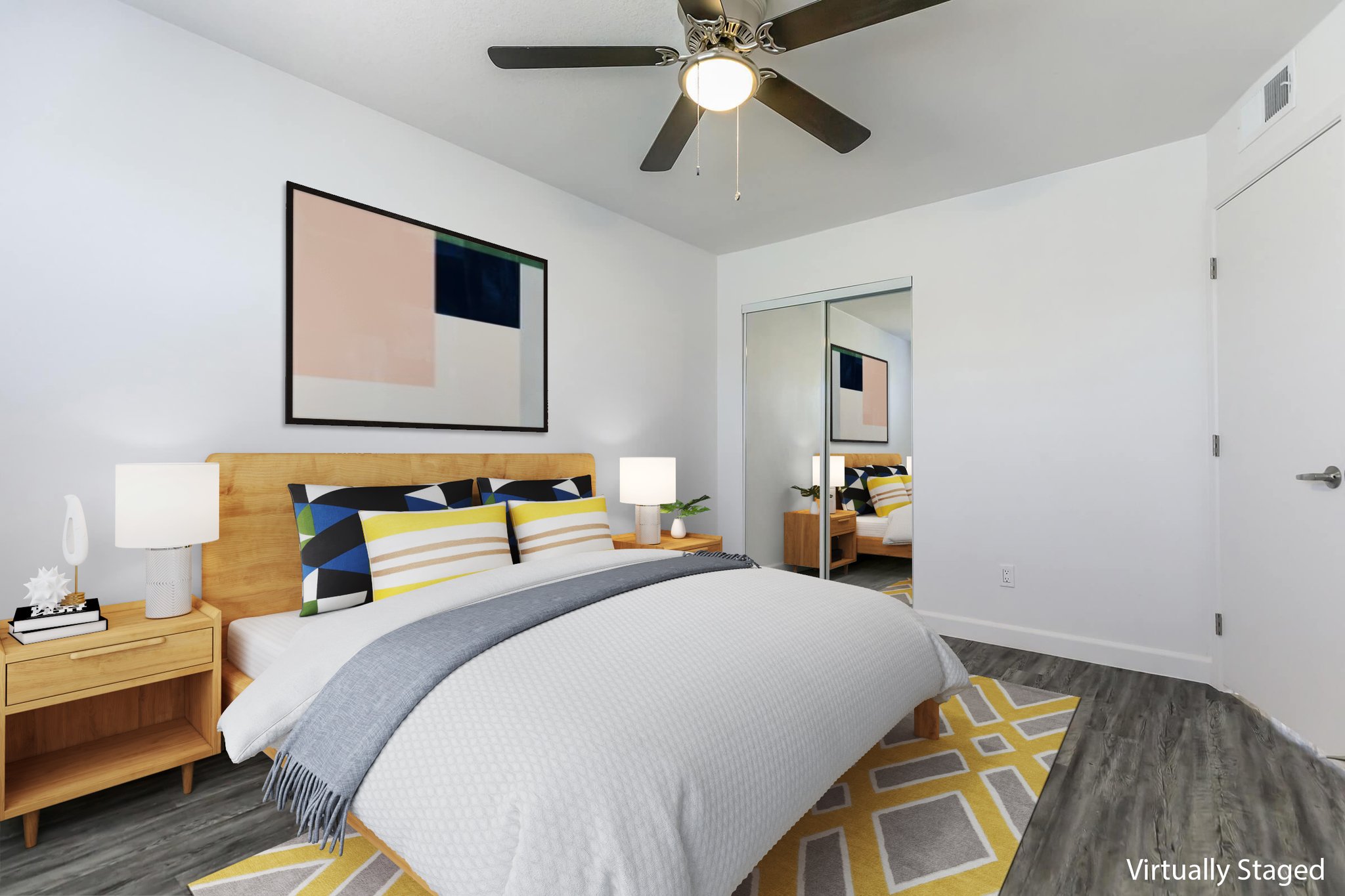 A modern bedroom featuring a double bed with yellow and gray bedding, two bedside tables with lamps, and a ceiling fan. A large piece of abstract art hangs above the bed, and a mirrored closet is visible on one side. The room has neutral walls and a geometric patterned rug on the floor.