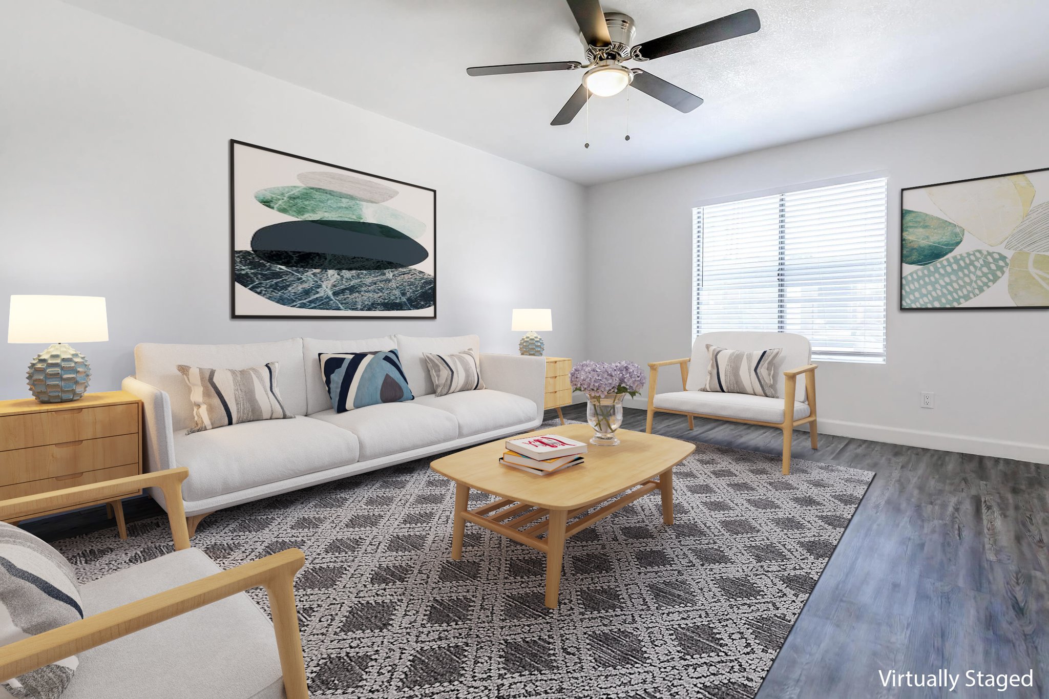 A bright and modern living room featuring a white sofa with decorative pillows, two light wooden chairs, and a coffee table with magazines. The space is adorned with abstract artwork on the walls, a ceiling fan, and soft lighting from two lamps. A bouquet of flowers adds a touch of color to the decor.
