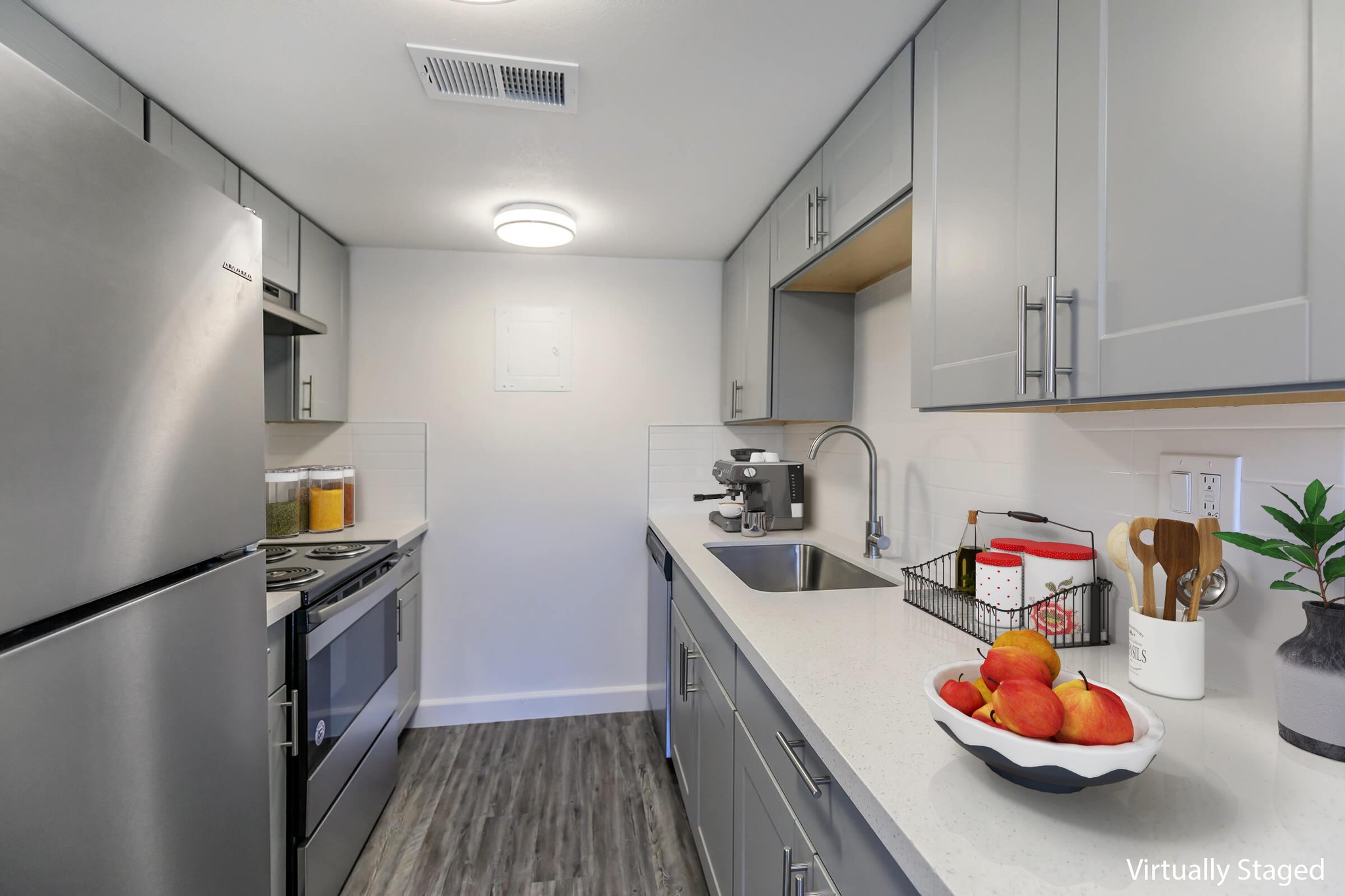 A modern kitchen featuring stainless steel appliances, including a refrigerator and oven. The cabinetry is gray with a sleek countertop and a sink. A small bowl of red apples is on the counter next to a coffee machine. Natural light brightens the space, which has wood-style flooring.
