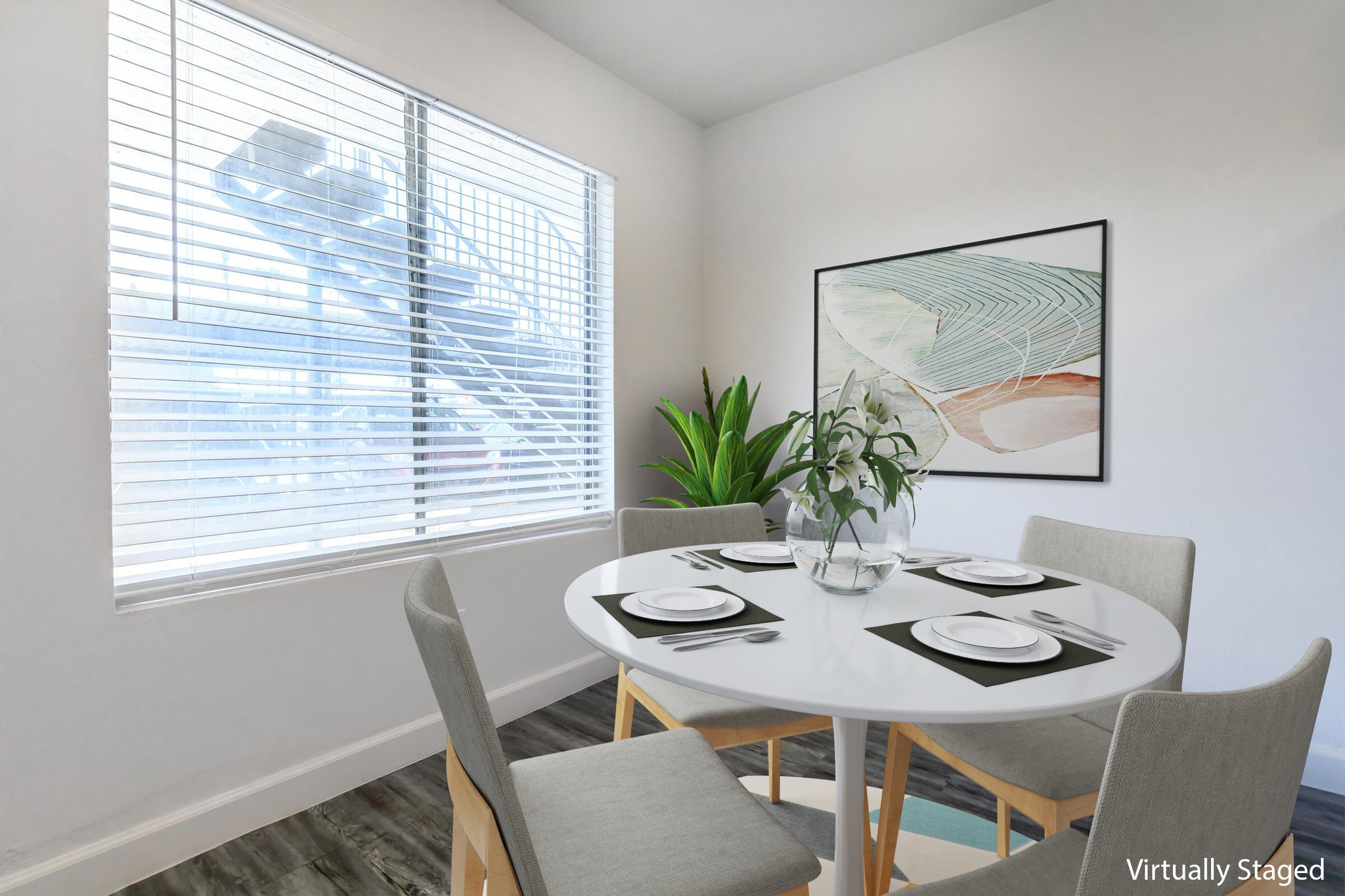 A cozy dining area featuring a round table set with plates and silverware. Four grey chairs surround the table. A large window with blinds allows natural light, and a potted plant adds a touch of greenery. A modern artwork hangs on the wall, enhancing the inviting atmosphere.