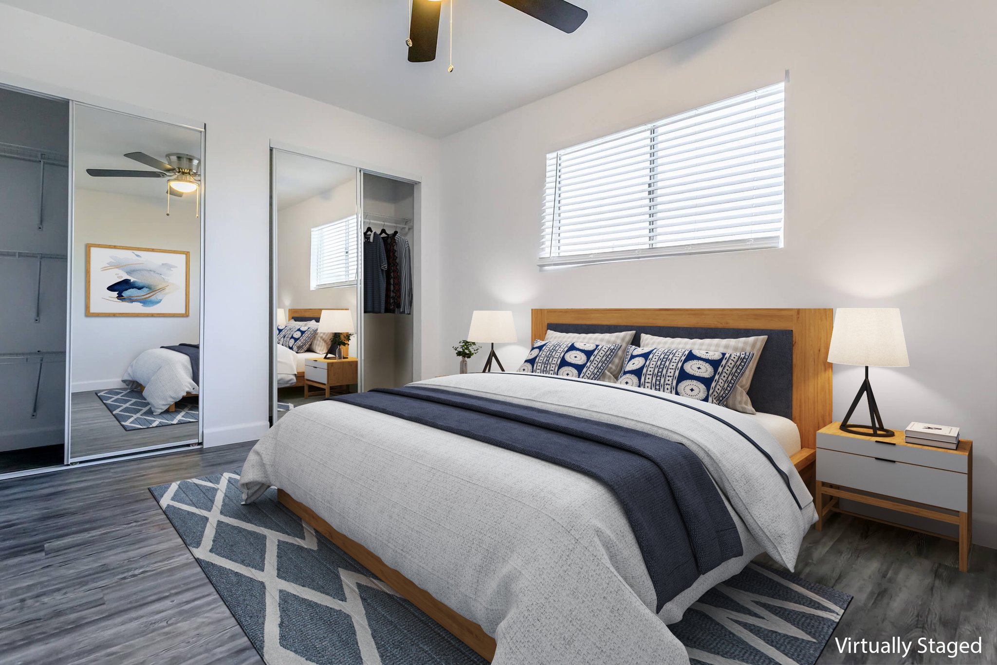 A modern bedroom featuring a large bed with blue and white bedding, nightstands with lamps, and a decorative area rug. A sliding mirror door showcases a closet, and a ceiling fan is visible. Natural light streams in through a window, enhancing the spacious and inviting atmosphere.