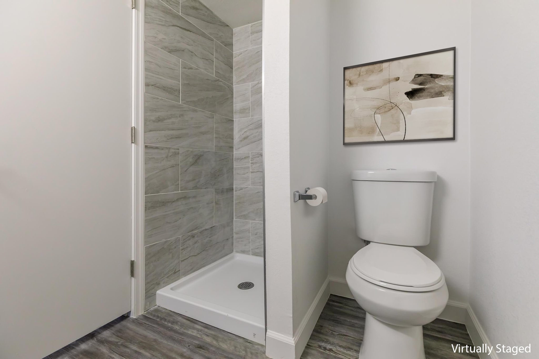 A modern bathroom featuring a walk-in shower with gray tiled walls, a white toilet, and a simple wall art piece. The floor has a wood-like finish, contributing to a clean and contemporary aesthetic. Light-colored walls enhance the spacious feel of the room.
