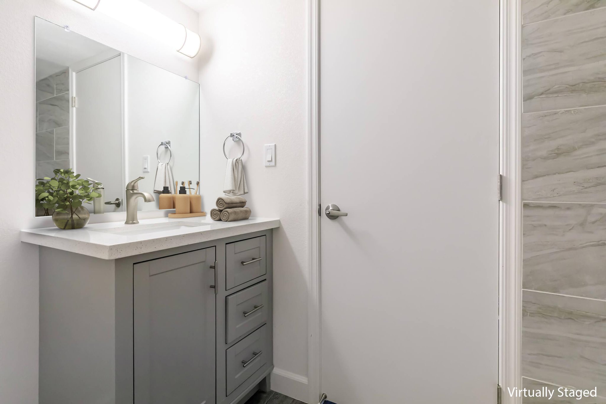 A modern bathroom featuring a light-colored countertop with a sink, gray cabinetry, and neatly rolled towels. A mirror above the sink reflects the room's simple decor. The wall opposite has a closed white door, with a small plant on the counter adding a touch of greenery. Soft lighting enhances the bright atmosphere.