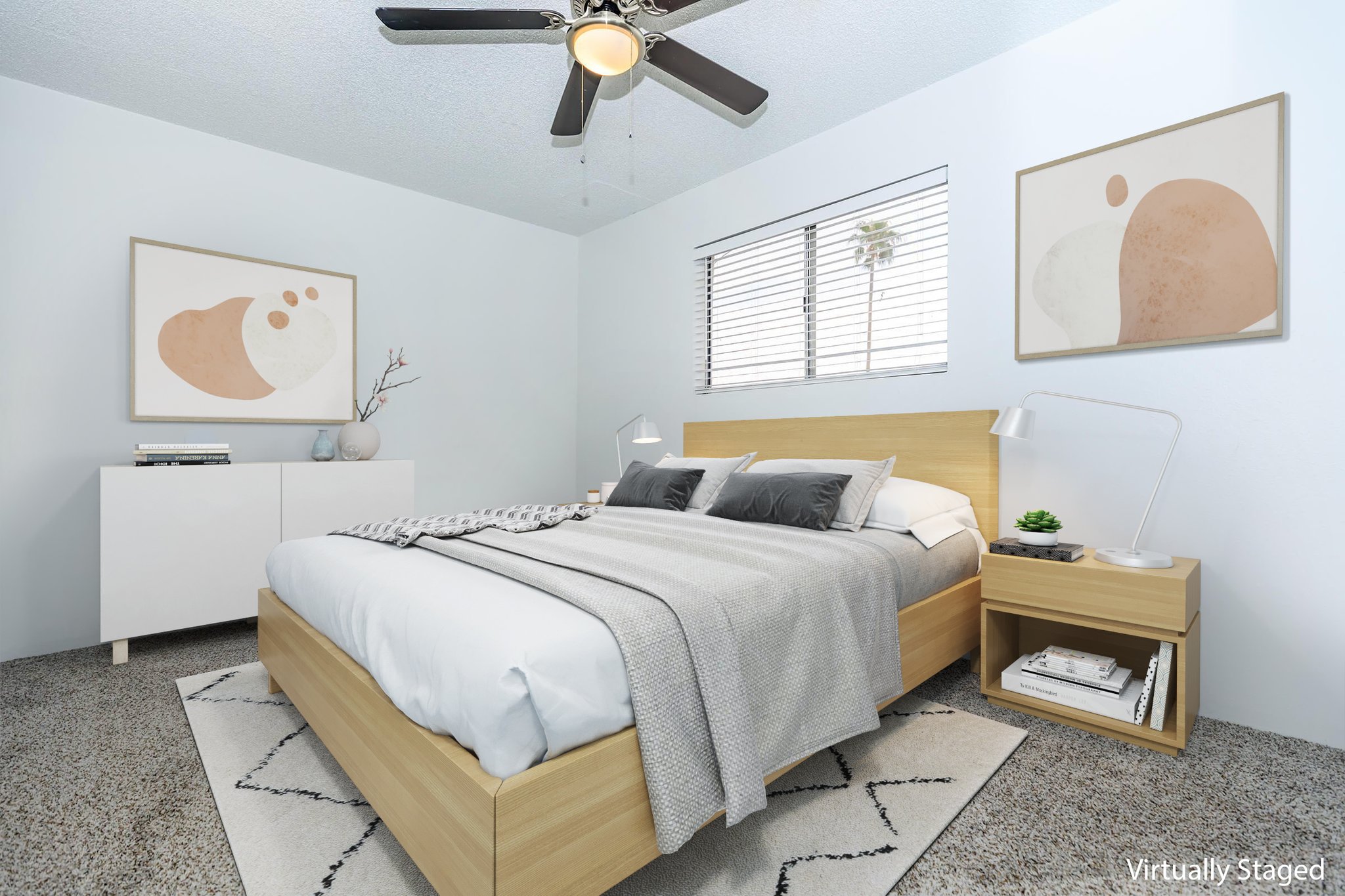 A modern, virtually staged bedroom featuring a wooden bed with gray bedding, a gray throw blanket, and decorative pillows. The room has light blue walls, two framed abstract artworks, a white dresser, a small nightstand with a lamp, and a ceiling fan. Natural light comes through the window, enhancing the cozy atmosphere.