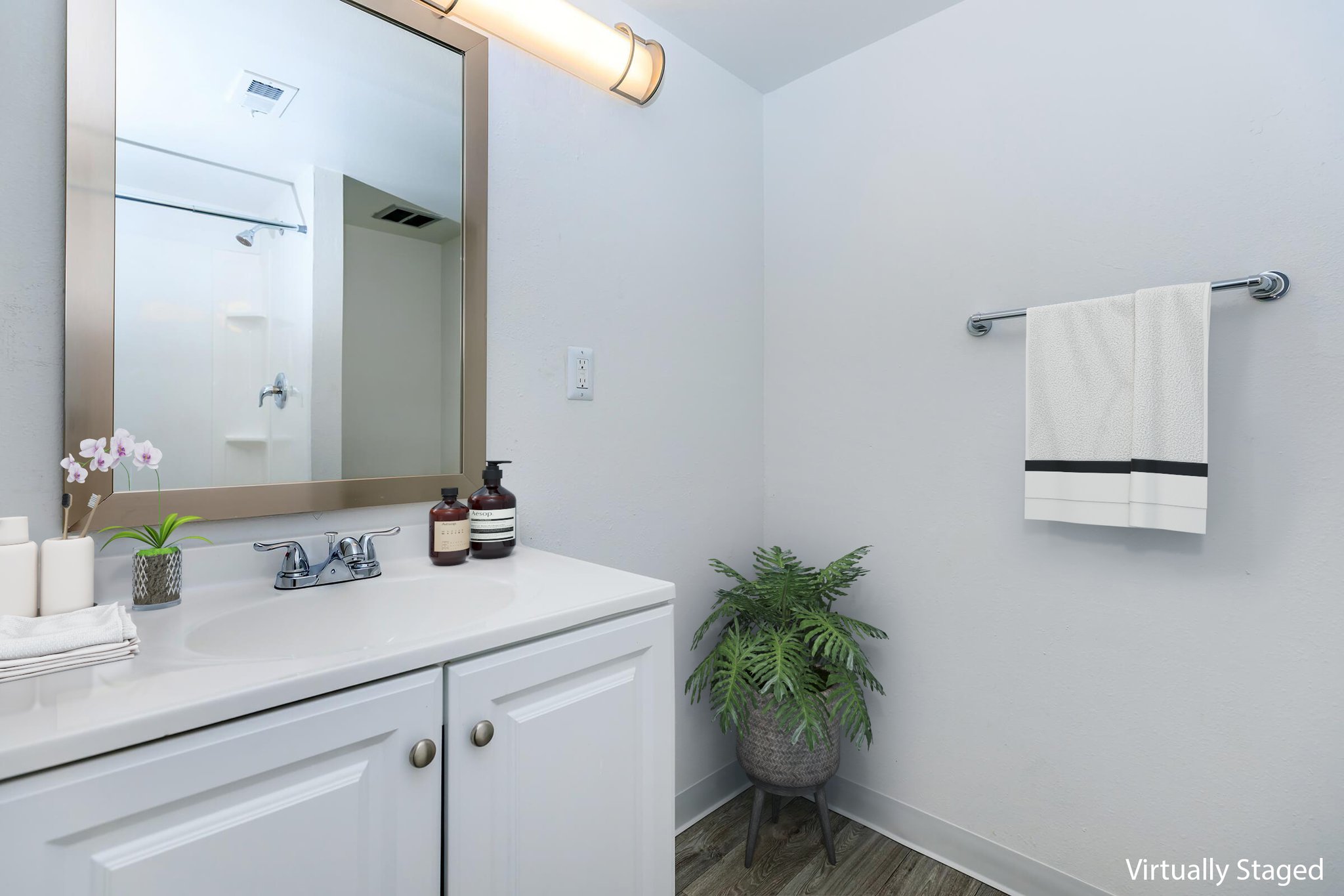 Modern bathroom featuring a white vanity with a sink, a large mirror illuminated by a wall-mounted light, and a towel rack with a neatly folded towel. Decorative plant and toiletries are displayed on the countertop, while the walls and flooring are in neutral tones, creating a clean and fresh atmosphere.
