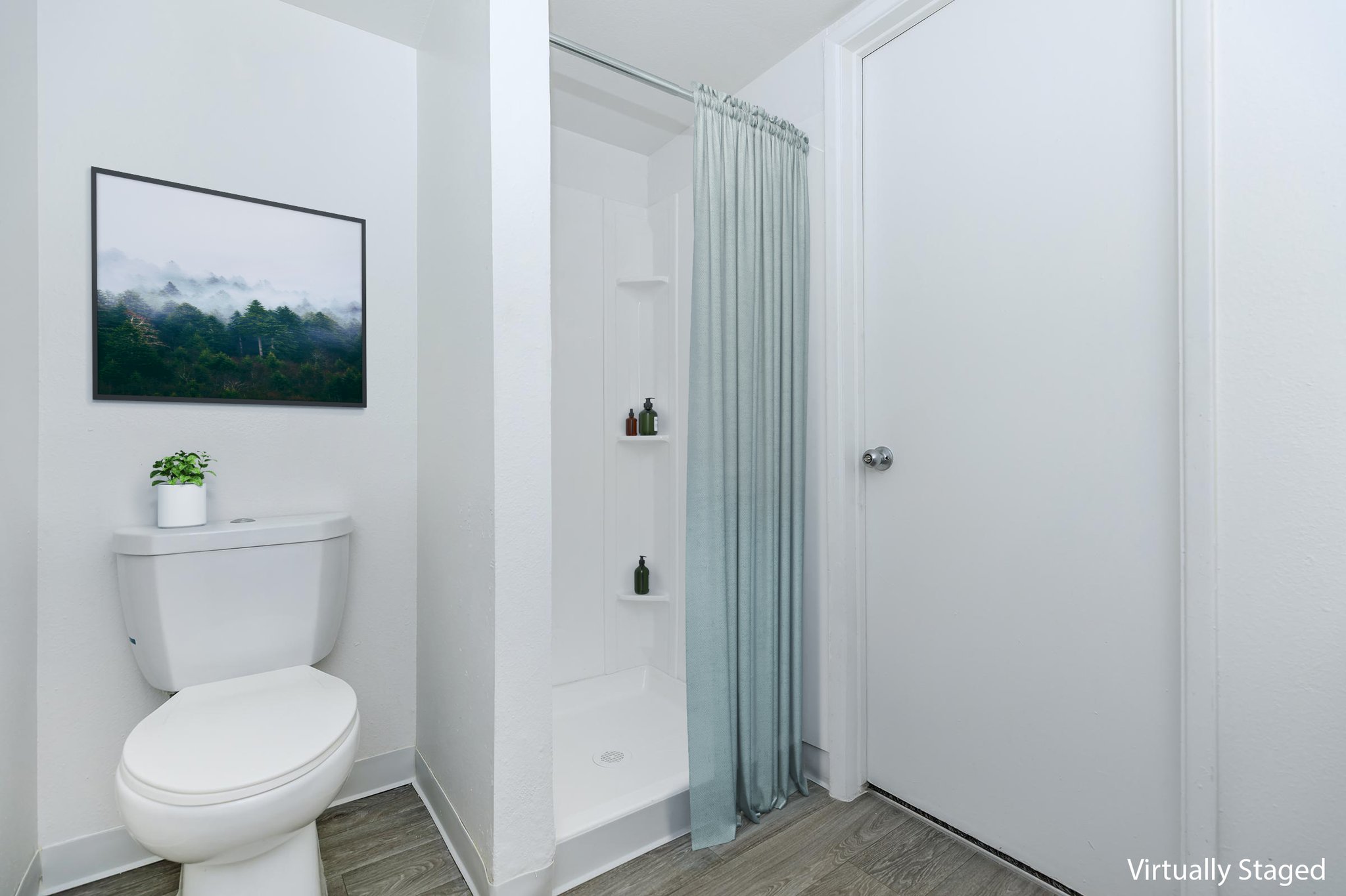 A modern bathroom featuring a white toilet, a shower with a light blue curtain, and a minimalist design. An artwork of a misty forest scene hangs on the wall, and there is a small potted plant on a shelf. The space has light-colored walls and wood-style flooring, creating a clean and inviting atmosphere.
