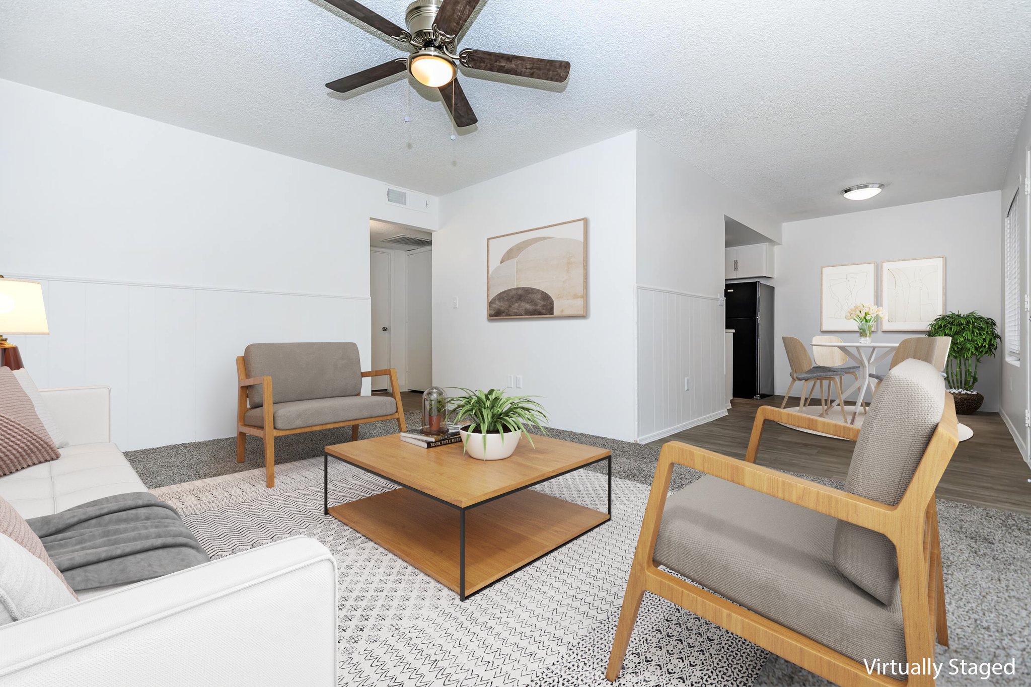 A cozy, modern living room featuring a light-colored sofa, two wooden armchairs, and a coffee table. The space has a textured rug and a ceiling fan, with a small plant on the table. The walls are painted white, and there are decorative pieces and a doorway leading to another room. Natural light brightens the area.