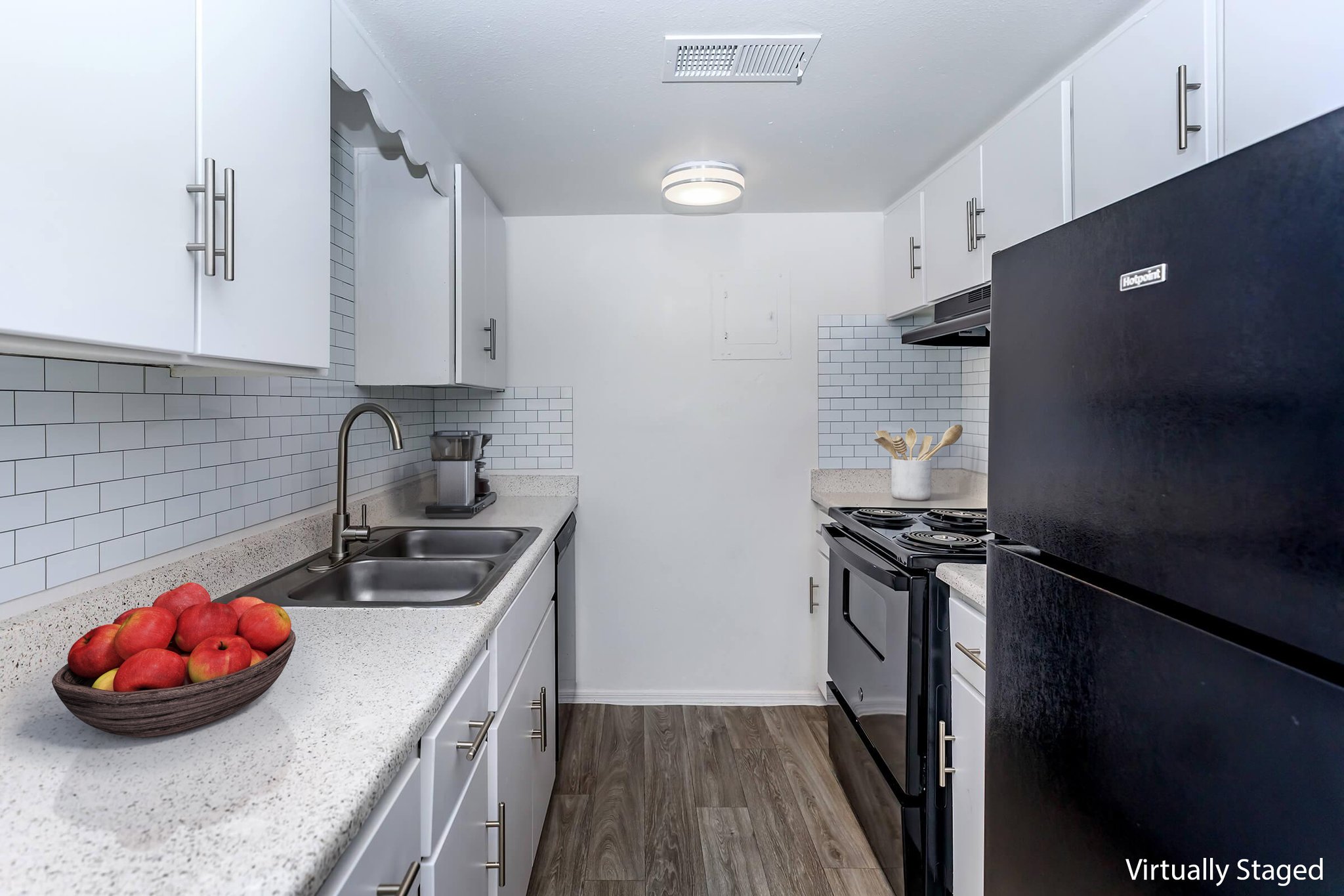 A modern kitchen featuring white cabinetry, a black refrigerator, and an electric stove. The countertops are light-colored with a bowl of red apples displayed. The backsplash consists of white subway tiles, and there is a stainless steel sink. Soft lighting illuminates the space, giving it a clean and inviting atmosphere.