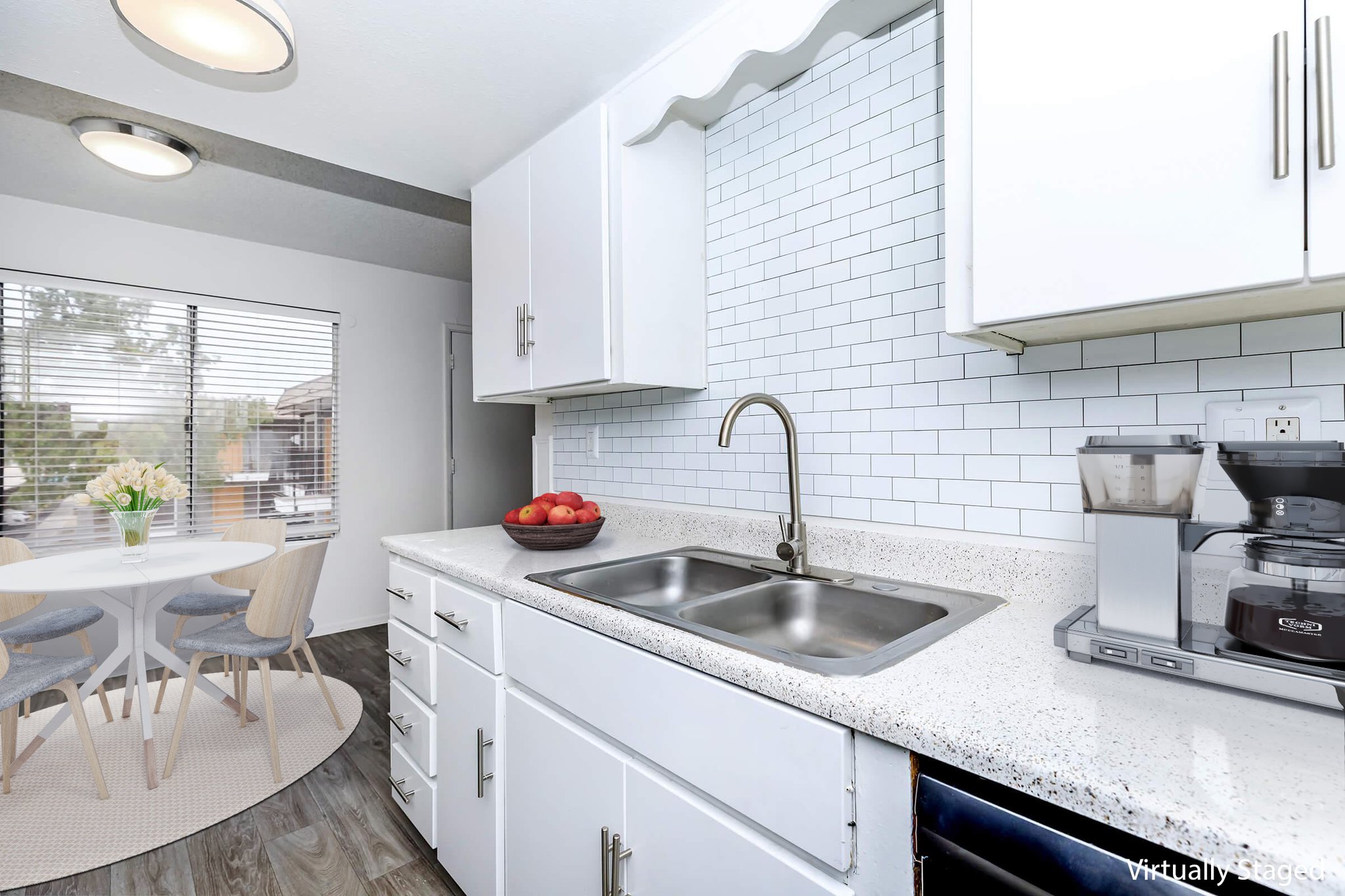 A bright, modern kitchen featuring white cabinets, a double sink, and a countertop with a speckled finish. A coffee maker and fruit bowl are visible, and a small dining area with a round table and light-colored chairs is seen through a window with blinds. Natural light illuminates the space.