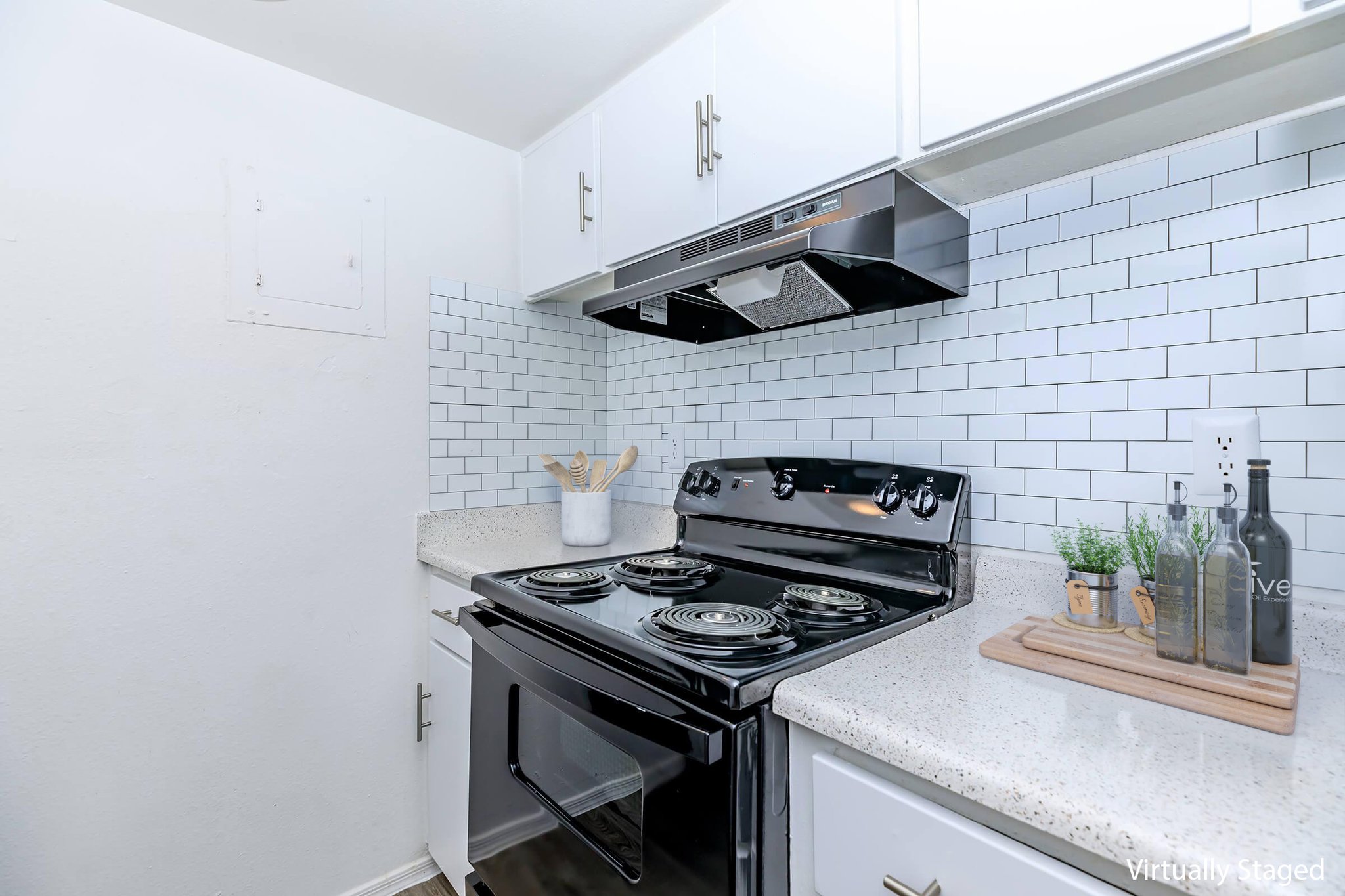 A modern kitchen featuring a black stove and oven, stainless steel range hood, and a light-colored countertop. The backsplash consists of white subway tiles. A wooden cutting board is placed on the counter along with decorative bottles and utensils. The cabinets are white, complementing the clean and contemporary design.