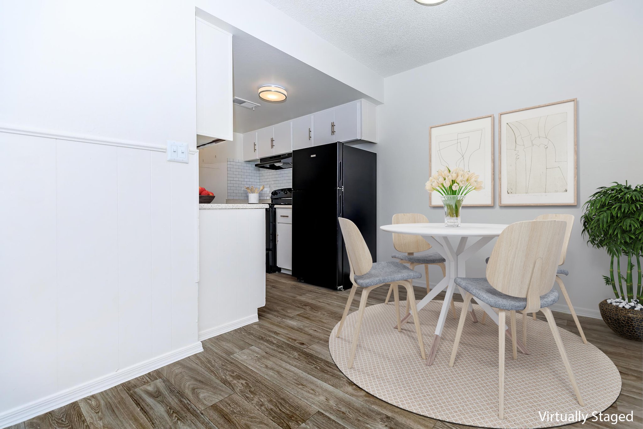 A bright dining area featuring a round white table with four wooden chairs, a decorative rug, and a vase of tulips. In the background, a black refrigerator and a white kitchen with modern appliances are visible. The space is tastefully staged with light-colored walls and art on display.