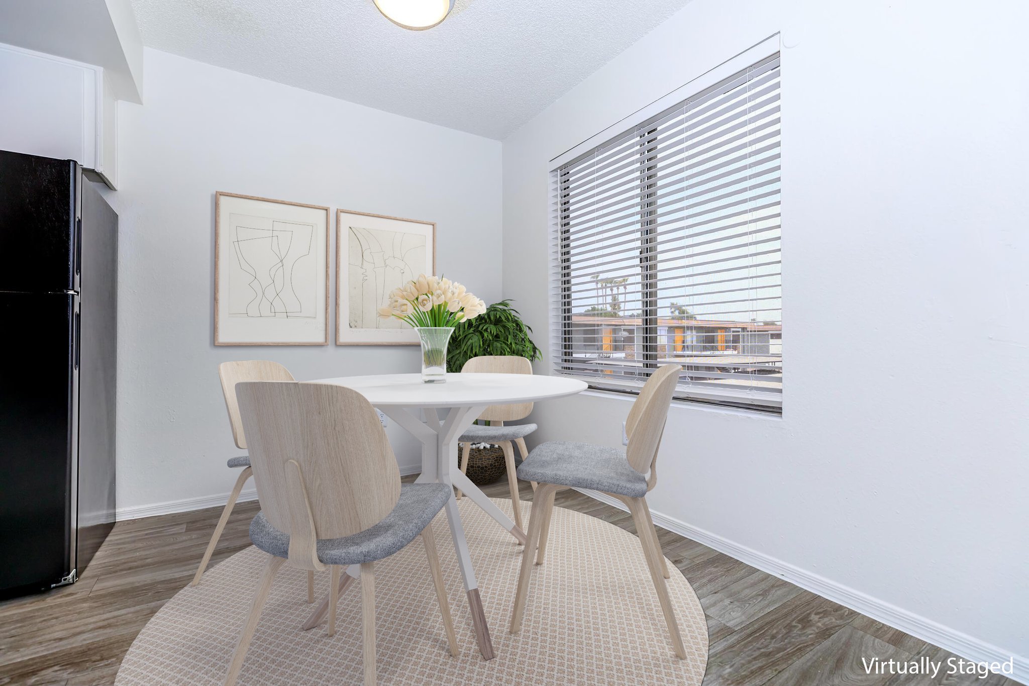 A modern dining area featuring a round white table with four light-colored chairs. A bouquet of flowers is placed in the center of the table. The room has large windows with blinds, allowing natural light to illuminate the space. Two framed artworks adorn the wall, and the floor has a light wooden finish.