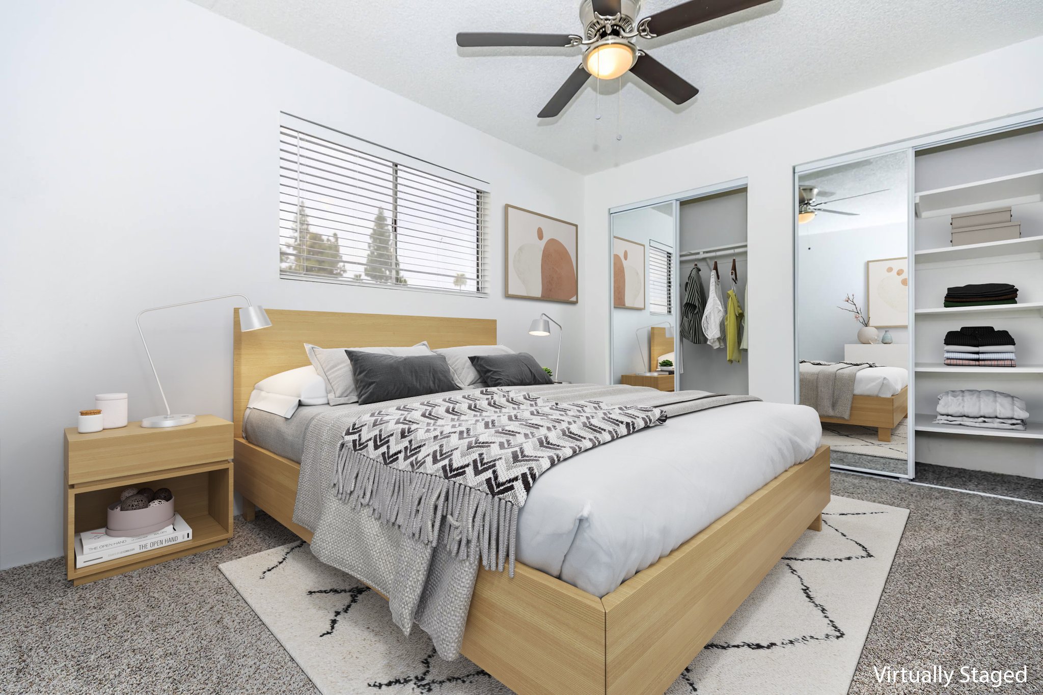 A modern bedroom featuring a wooden bed with gray and white bedding. There is a nightstand with a lamp beside the bed. A mirrored closet is visible, showing organized items. The room has a ceiling fan and a light-colored rug with a geometric pattern. Natural light comes through the window with simple wall art above the bed.