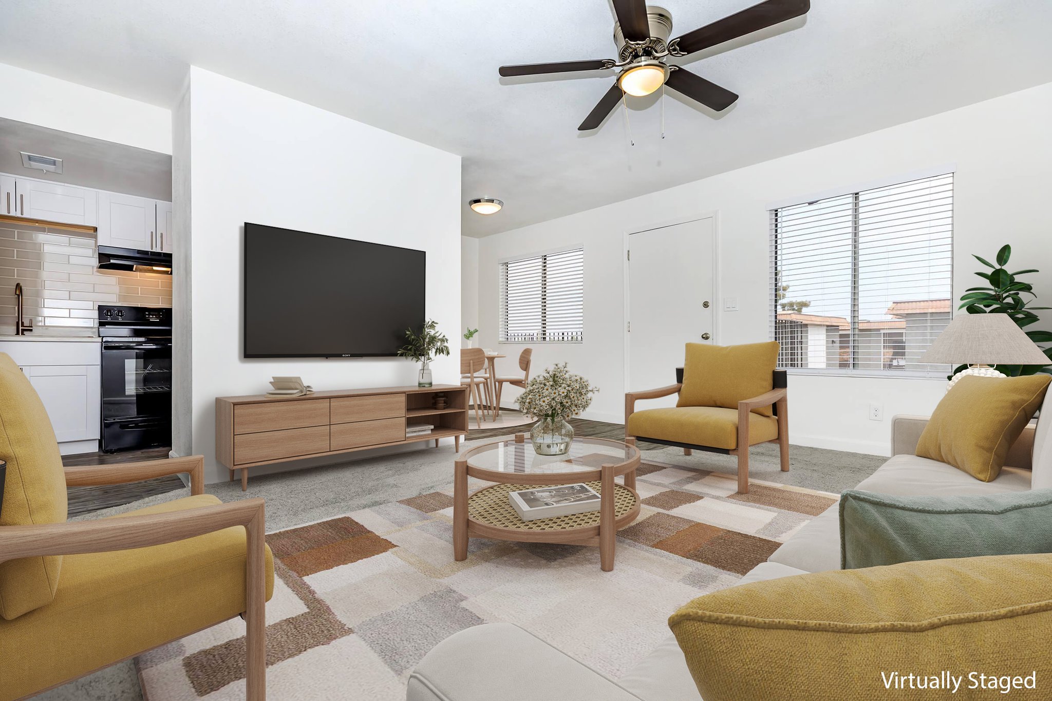 A modern living room featuring a comfortable seating arrangement with yellow and beige chairs, a neutral sofa, and a round coffee table. The space contains a large flat-screen TV mounted on the wall, a window with blinds, and a small dining area in the background, all designed in a bright and inviting style.
