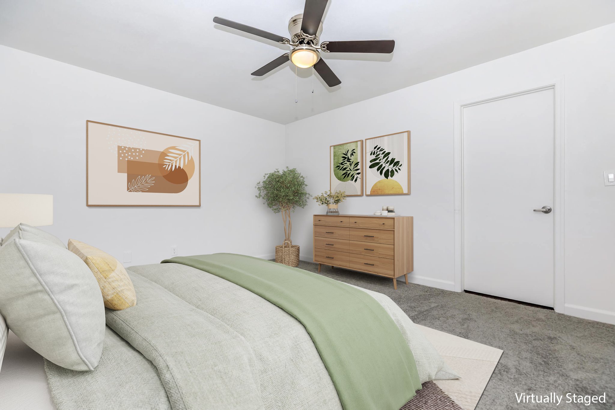 A modern, virtually staged bedroom featuring a light gray bed with green and beige bedding. Two framed botanical art pieces on the white walls, a leafy plant in a pot beside a wooden dresser, and a ceiling fan above. The room has a neutral carpet and a closed white door in the background.