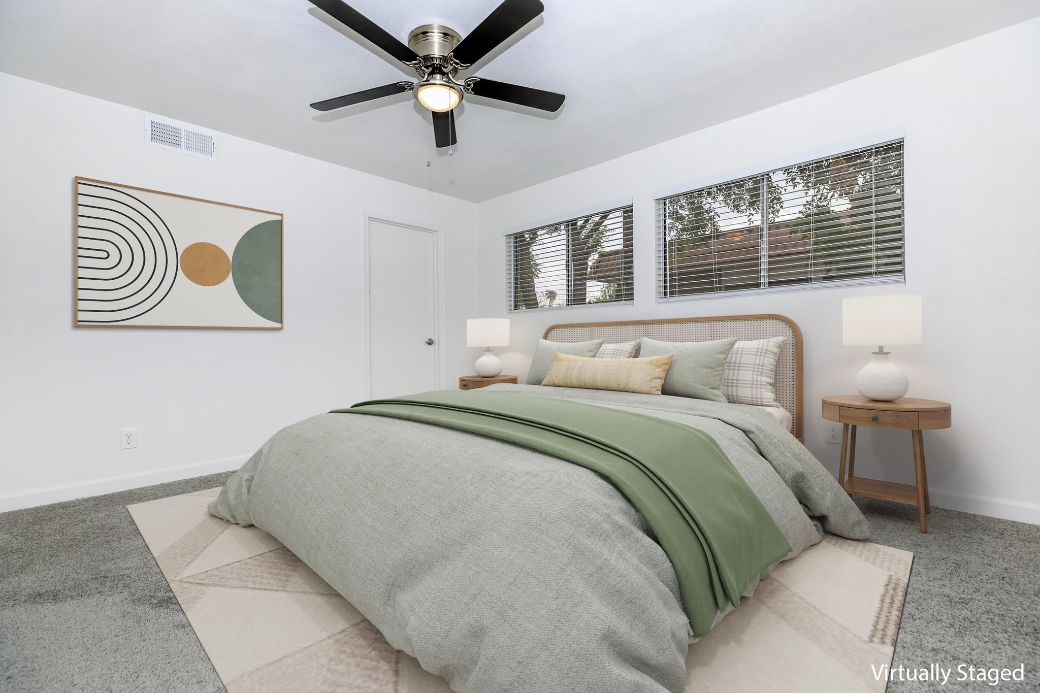 A modern bedroom featuring a queen-sized bed with gray and green bedding, two bedside lamps on circular tables, and a large wall art piece. The room includes a ceiling fan, large windows with blinds, and light-colored walls and carpet, creating a bright and inviting atmosphere. Virtually staged for decor illustration.