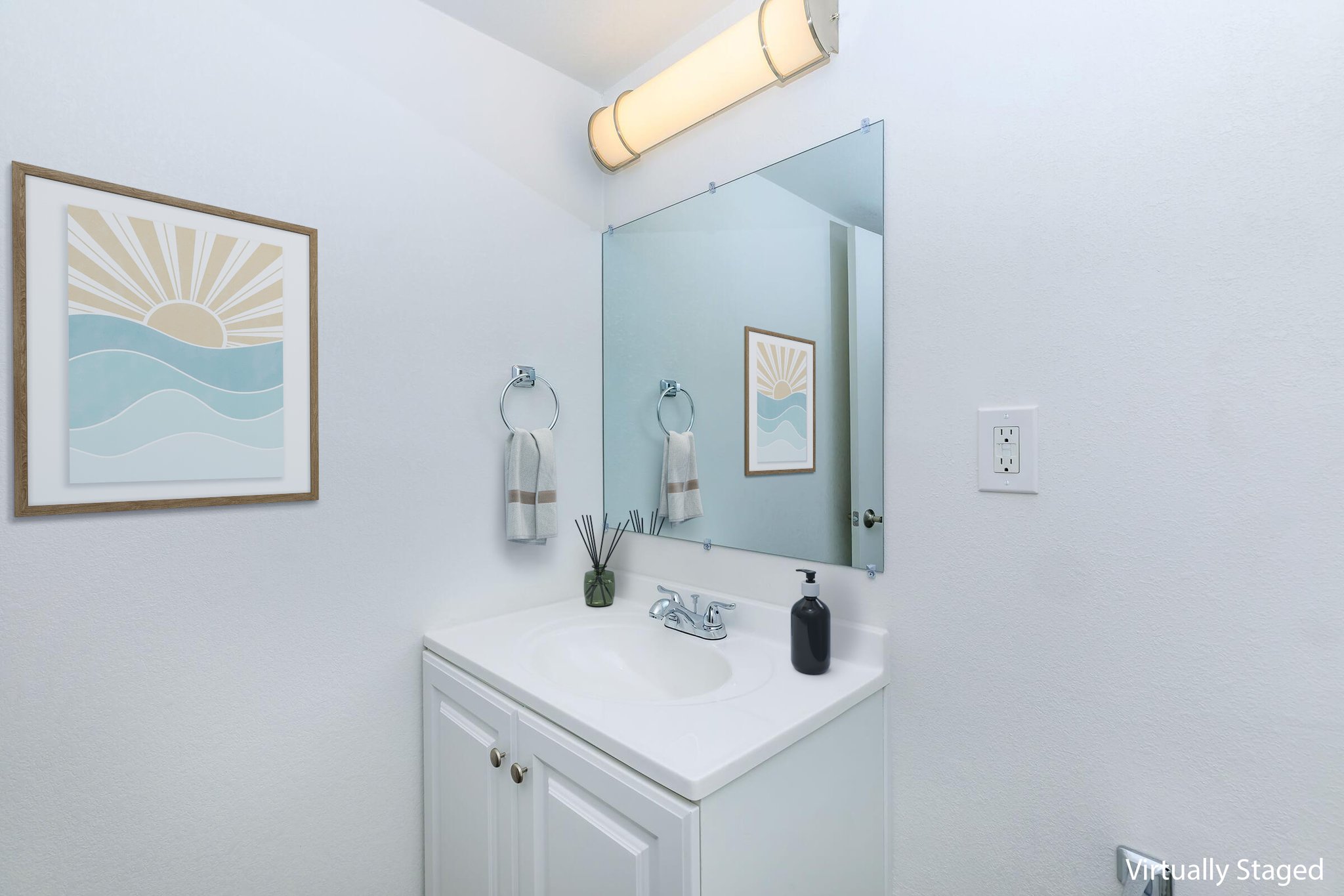 A clean, modern bathroom featuring a white vanity with a sink, a large mirror, and a framed wall art depicting waves and a sun. There are two hand towels on towel rings, decorative items on the countertop, and neutral-colored walls. The lighting is bright and inviting.