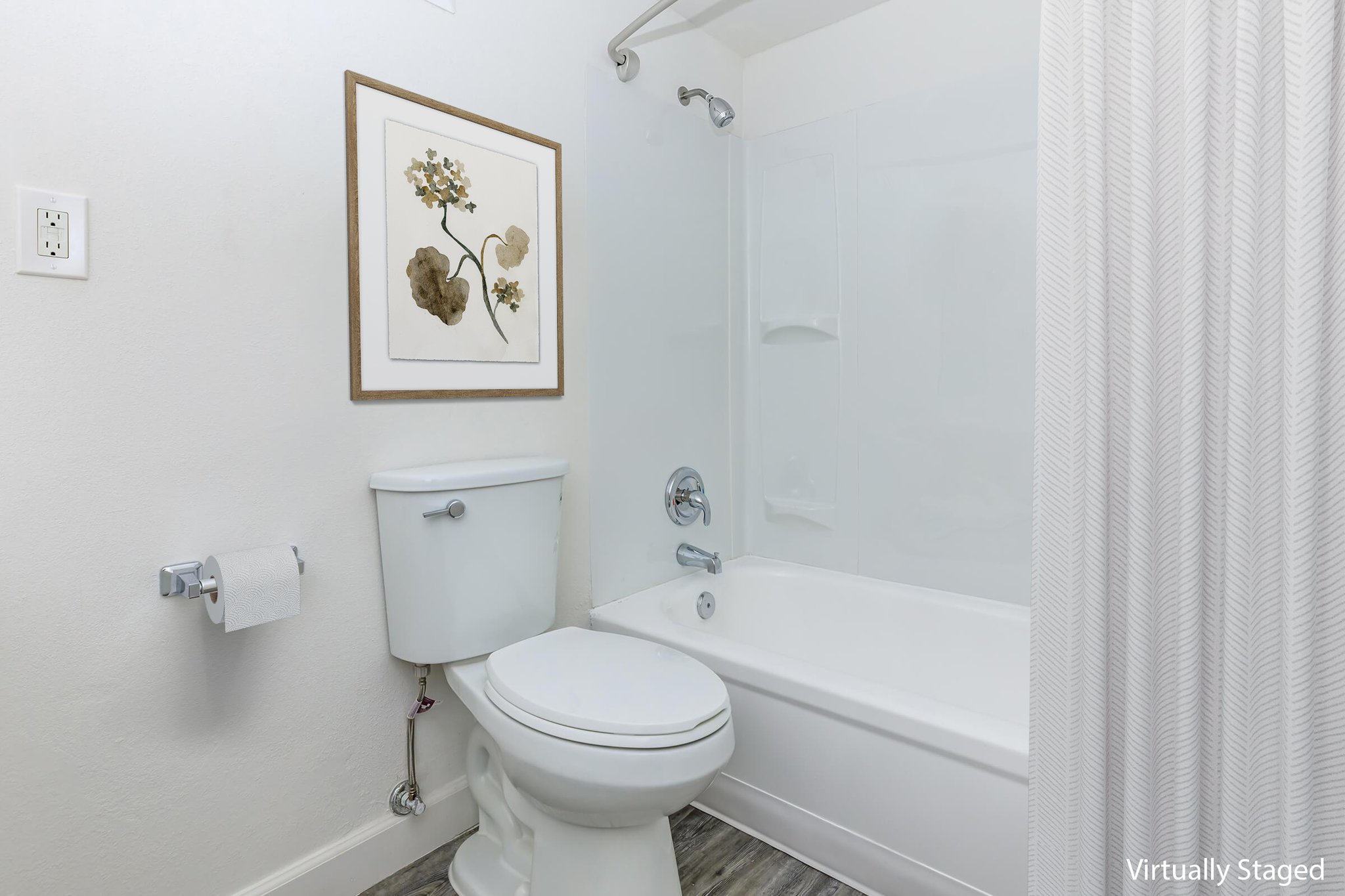 A clean, modern bathroom featuring a white bathtub with a shower, a toilet, and a framed floral artwork on the wall. The decor includes light-colored walls and a textured shower curtain, with a toilet paper holder on the side. The flooring is a wood-like material, adding a warm touch to the space.